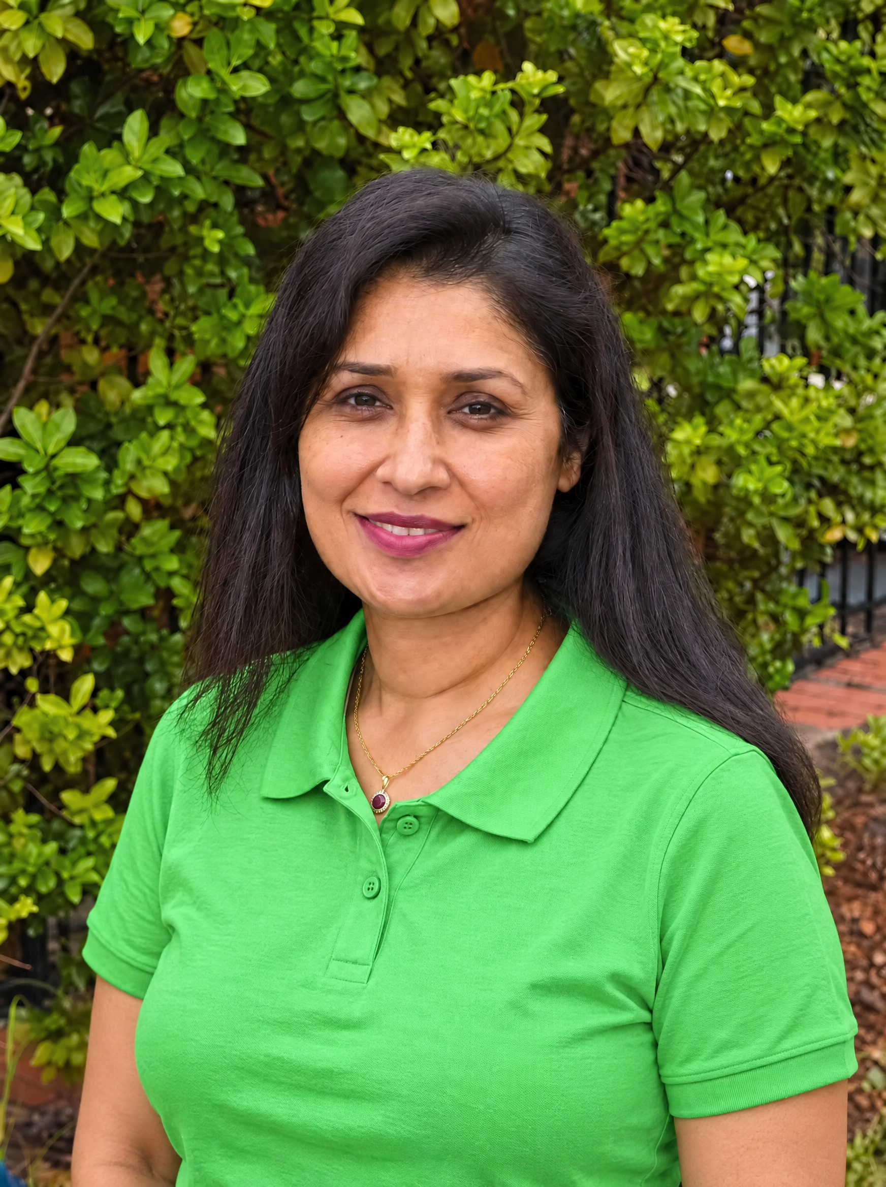 A woman with long black hair wearing a green shirt, black jacket, and a gold necklace, smiling outdoors with green foliage in the background.