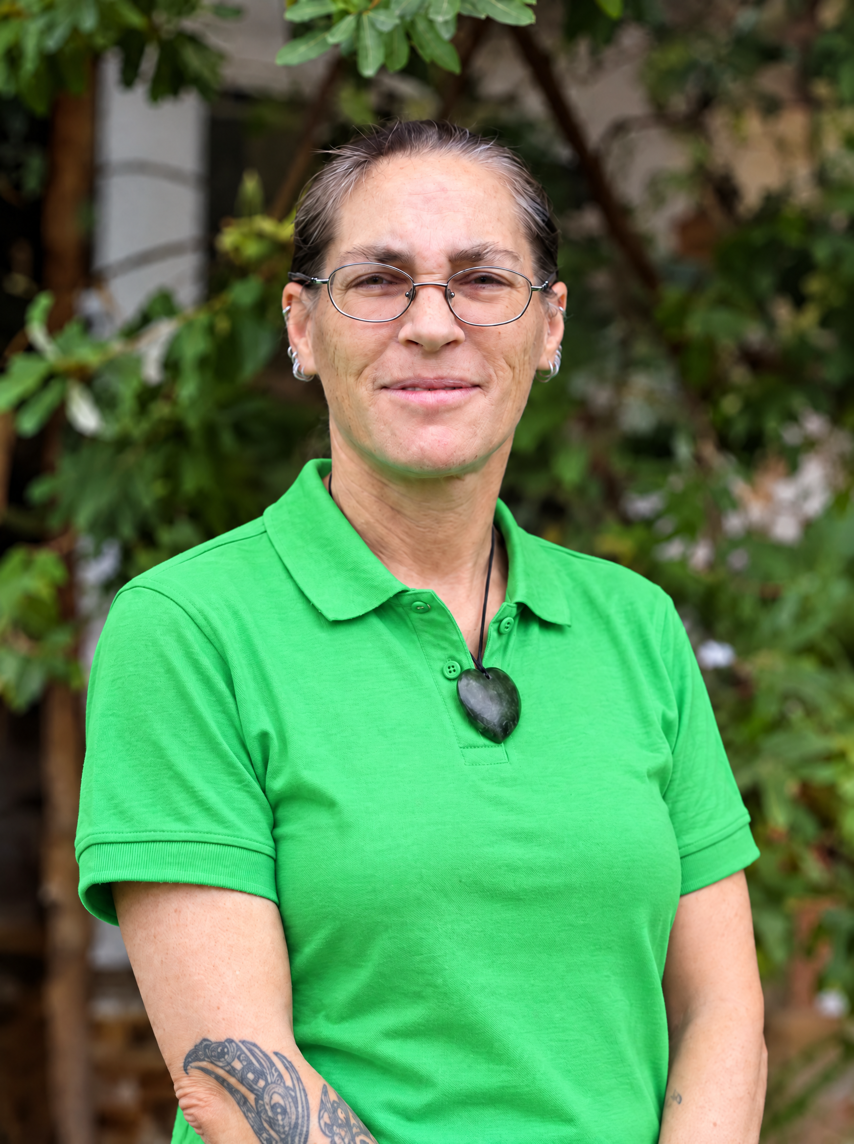 Person wearing a green shirt stands outdoors with trees and a blue canopy in the background.