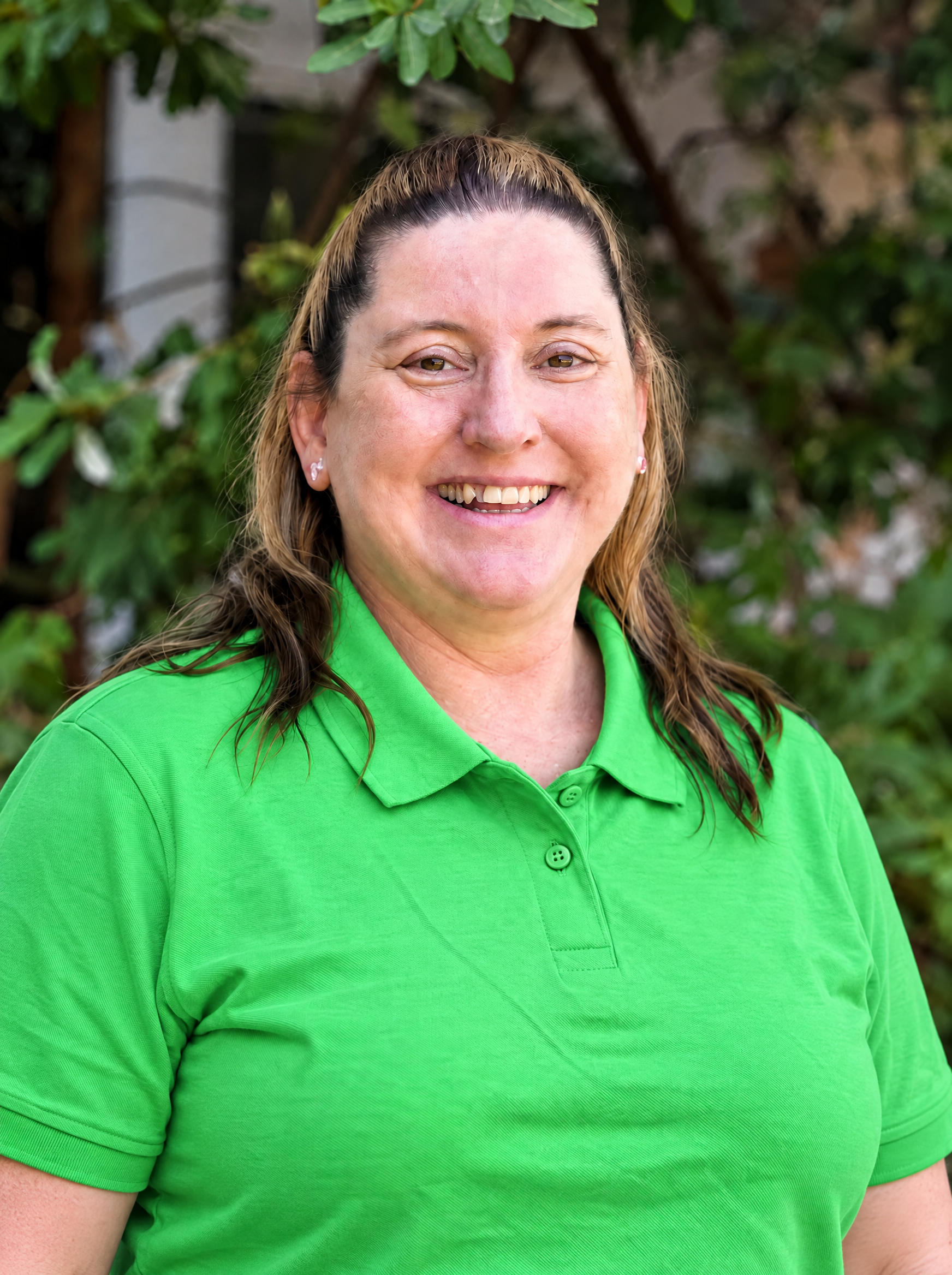 A woman smiling, wearing a green polo shirt and black jacket, standing in a classroom with educational decorations on the wall.