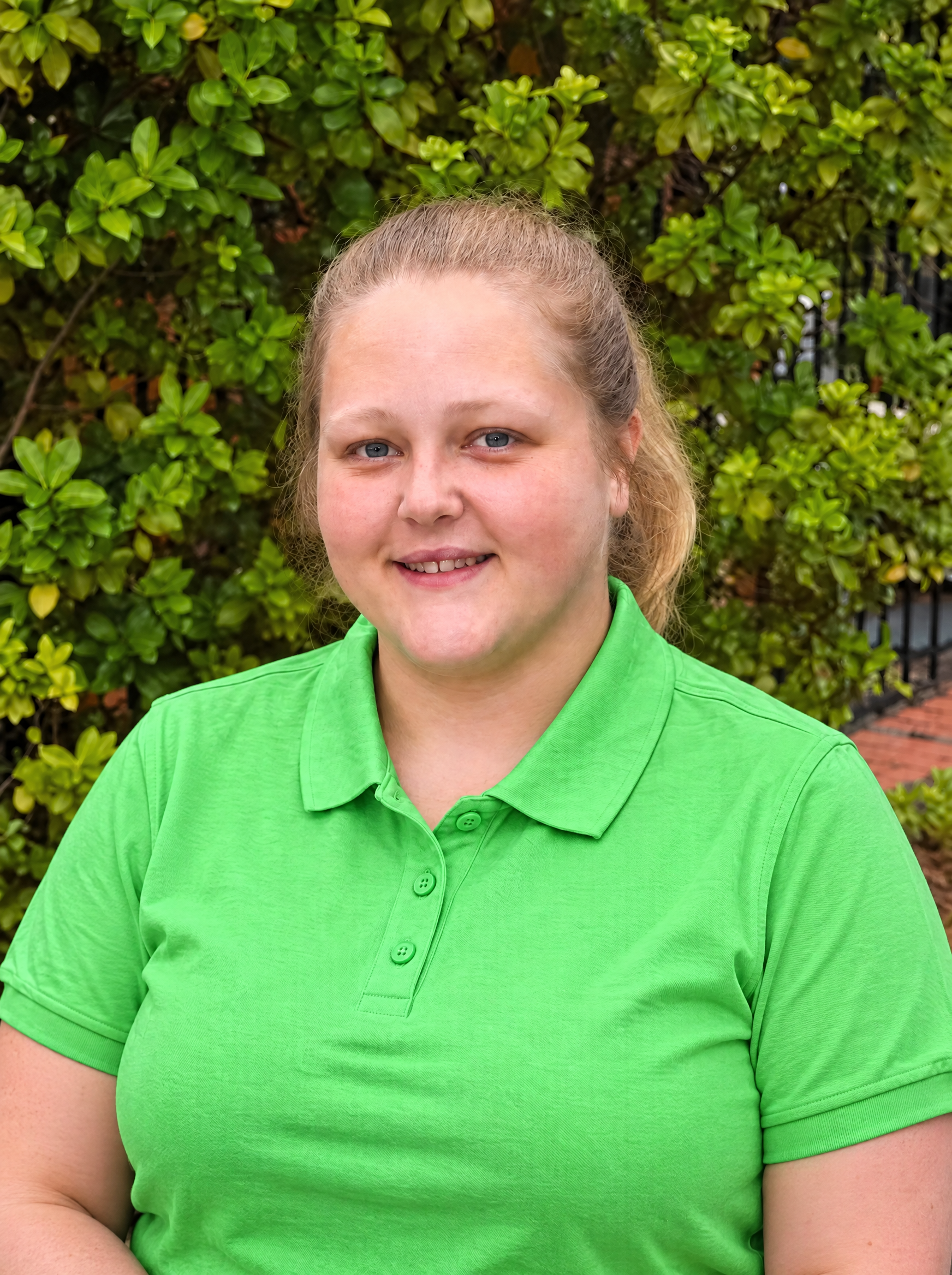 A woman with blonde hair tied back, wearing a green polo shirt and a black Adidas jacket, standing outdoors in front of green foliage.