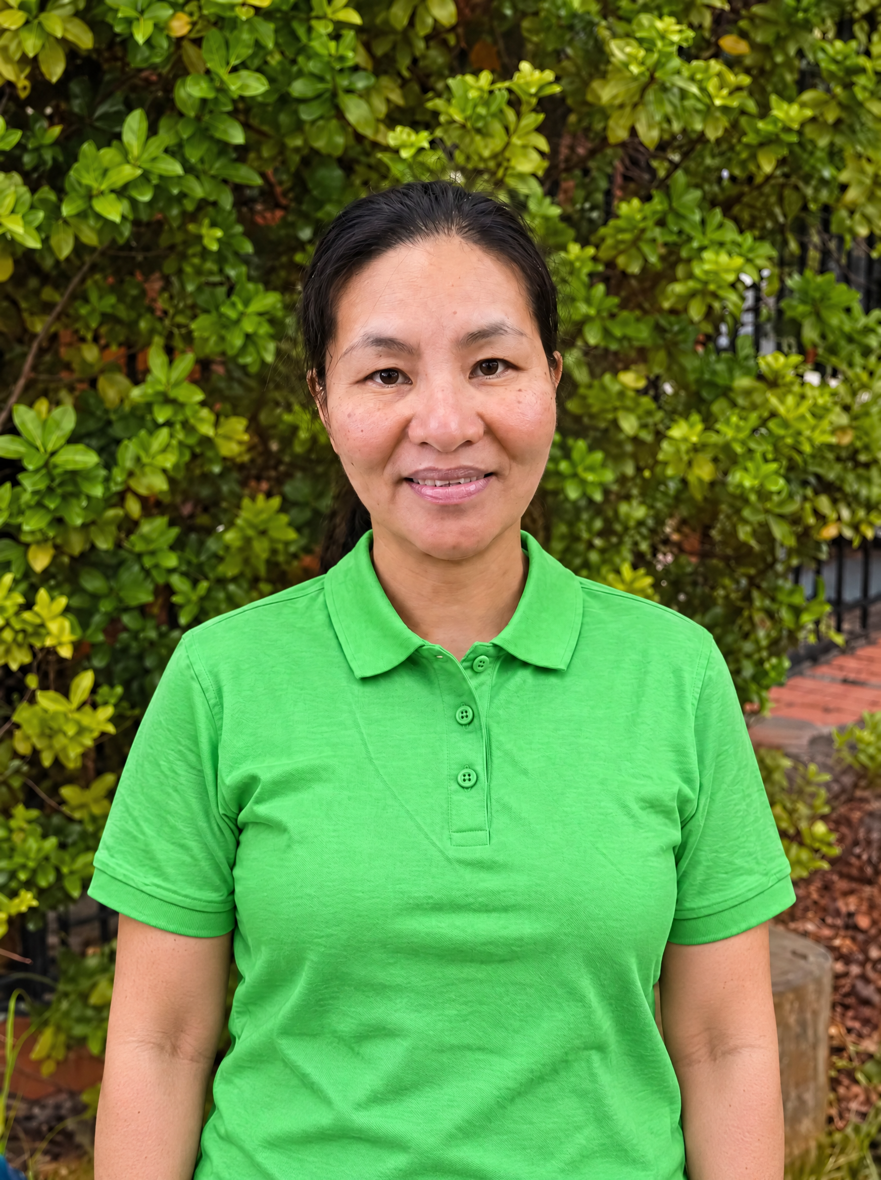 A woman with dark hair in a ponytail, wearing a bright green polo shirt over a black long-sleeve shirt, standing against a brick wall.