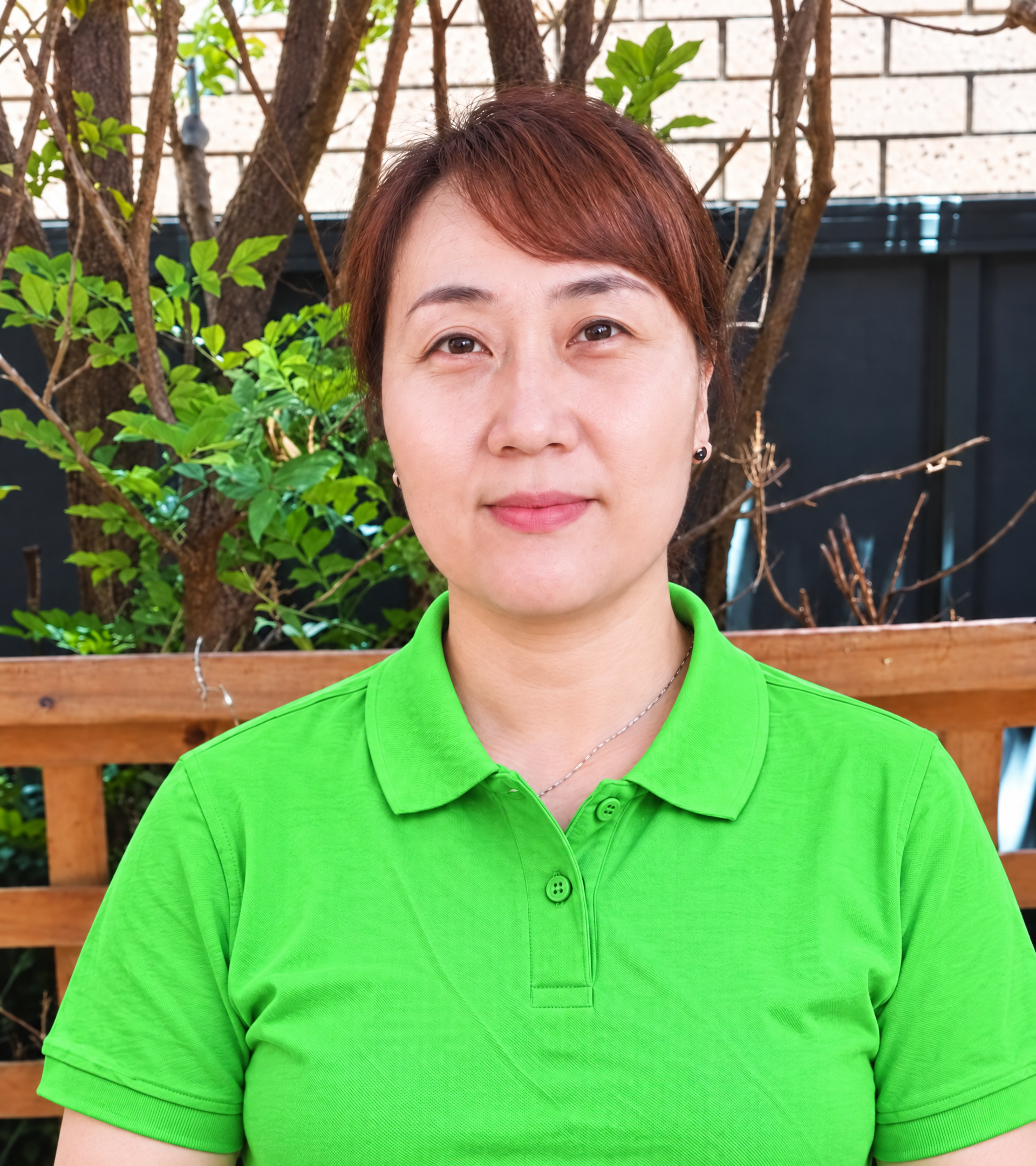 A woman with long black hair wearing a green shirt, black jacket, and a gold necklace, smiling outdoors with green foliage in the background.