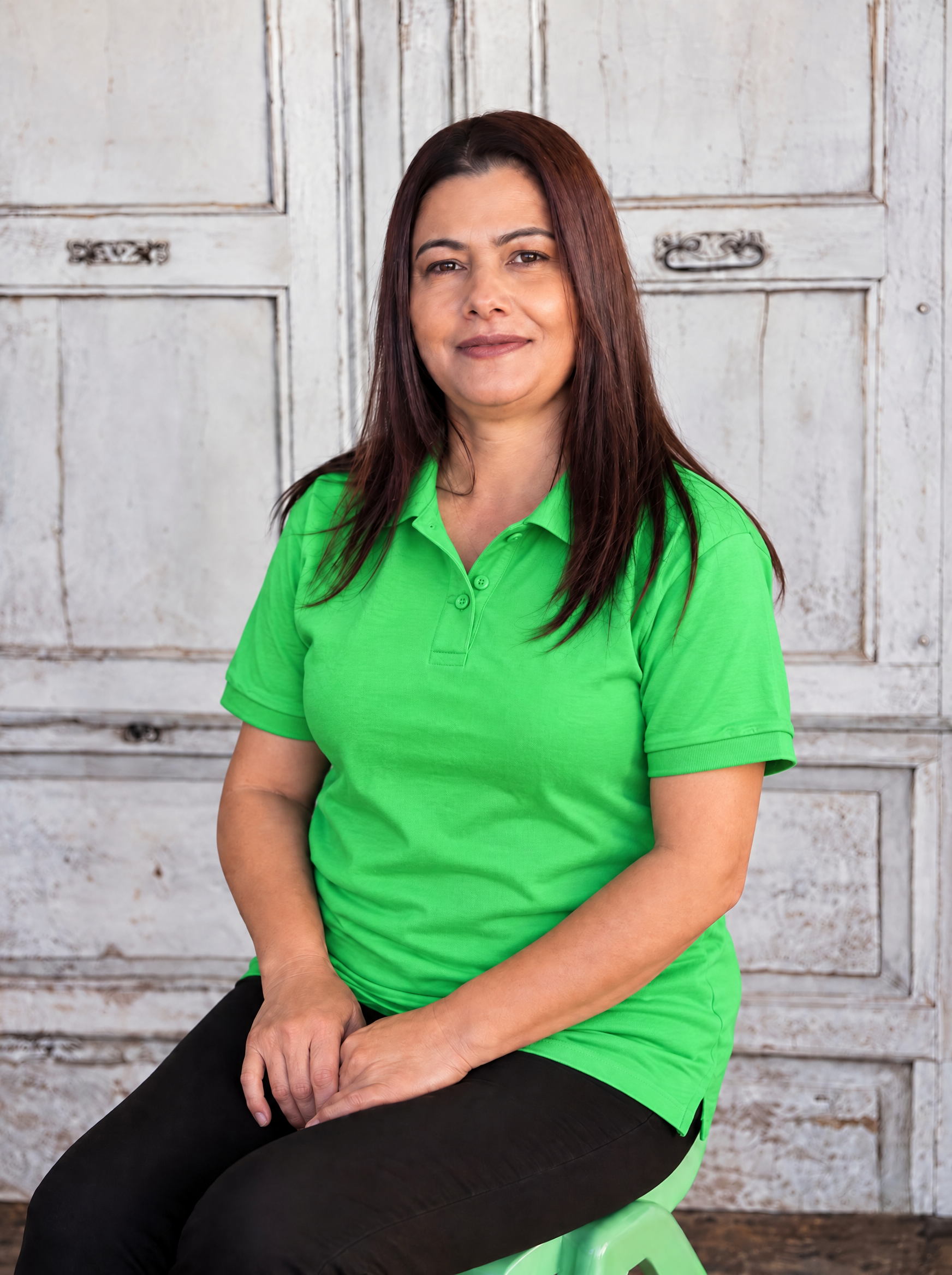 A woman with long black hair wearing a green shirt, black jacket, and a gold necklace, smiling outdoors with green foliage in the background.