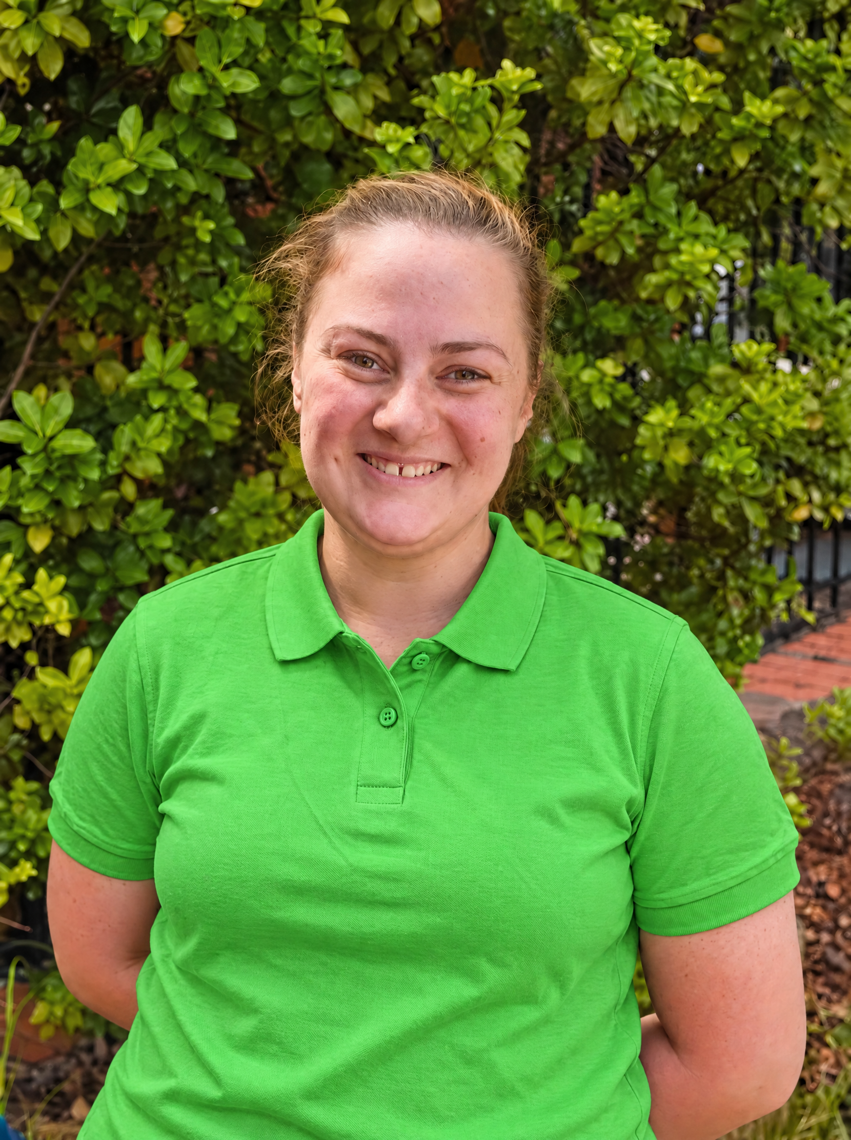 A smiling woman with light skin and red hair standing outdoors in front of a leafy green bush on a sunny day.