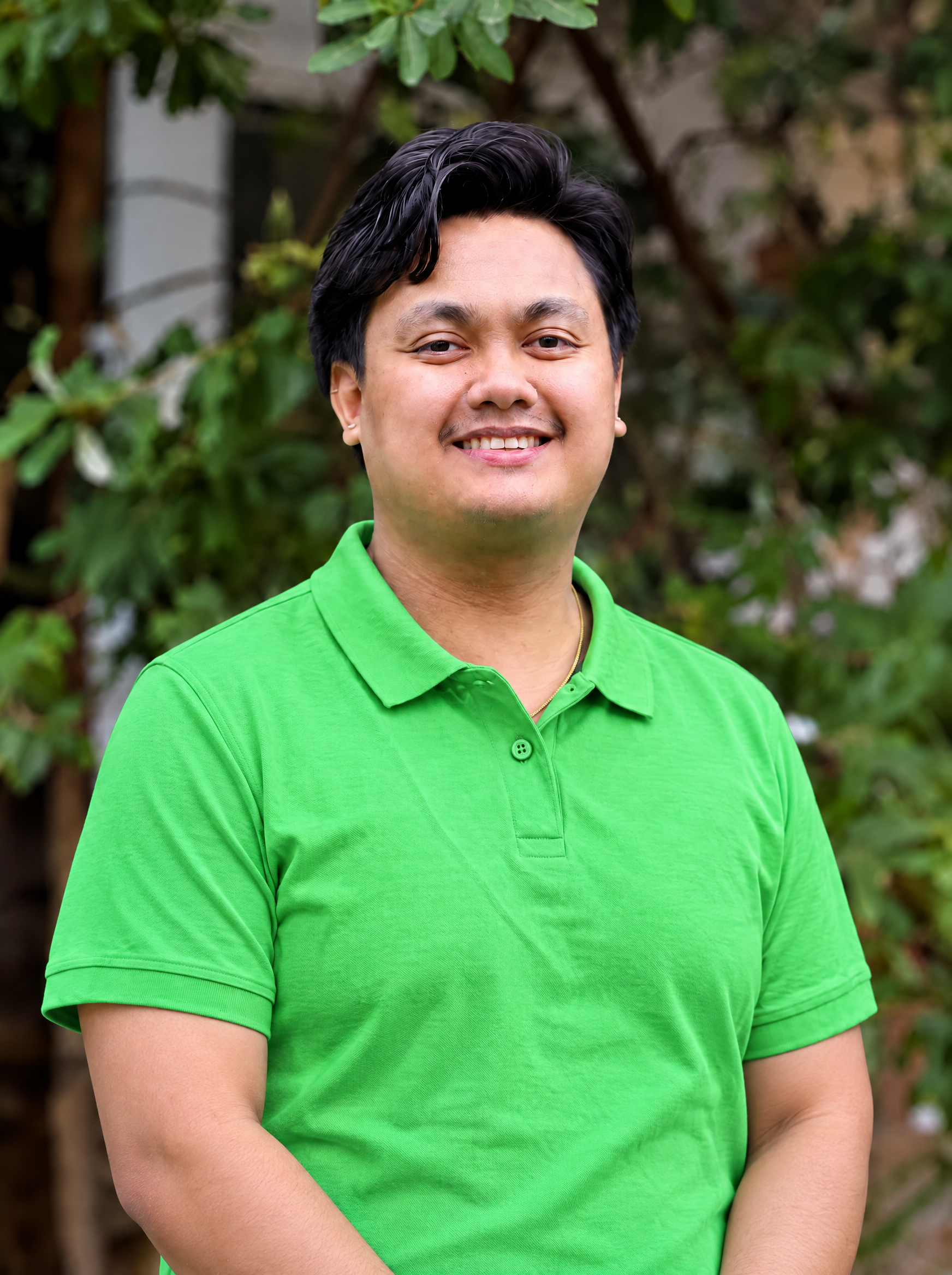 A young man with short black hair and a slight smile, wearing a bright green polo shirt over a black long-sleeve shirt, standing in front of a wall decorated with numerous picture frames and children's drawings.