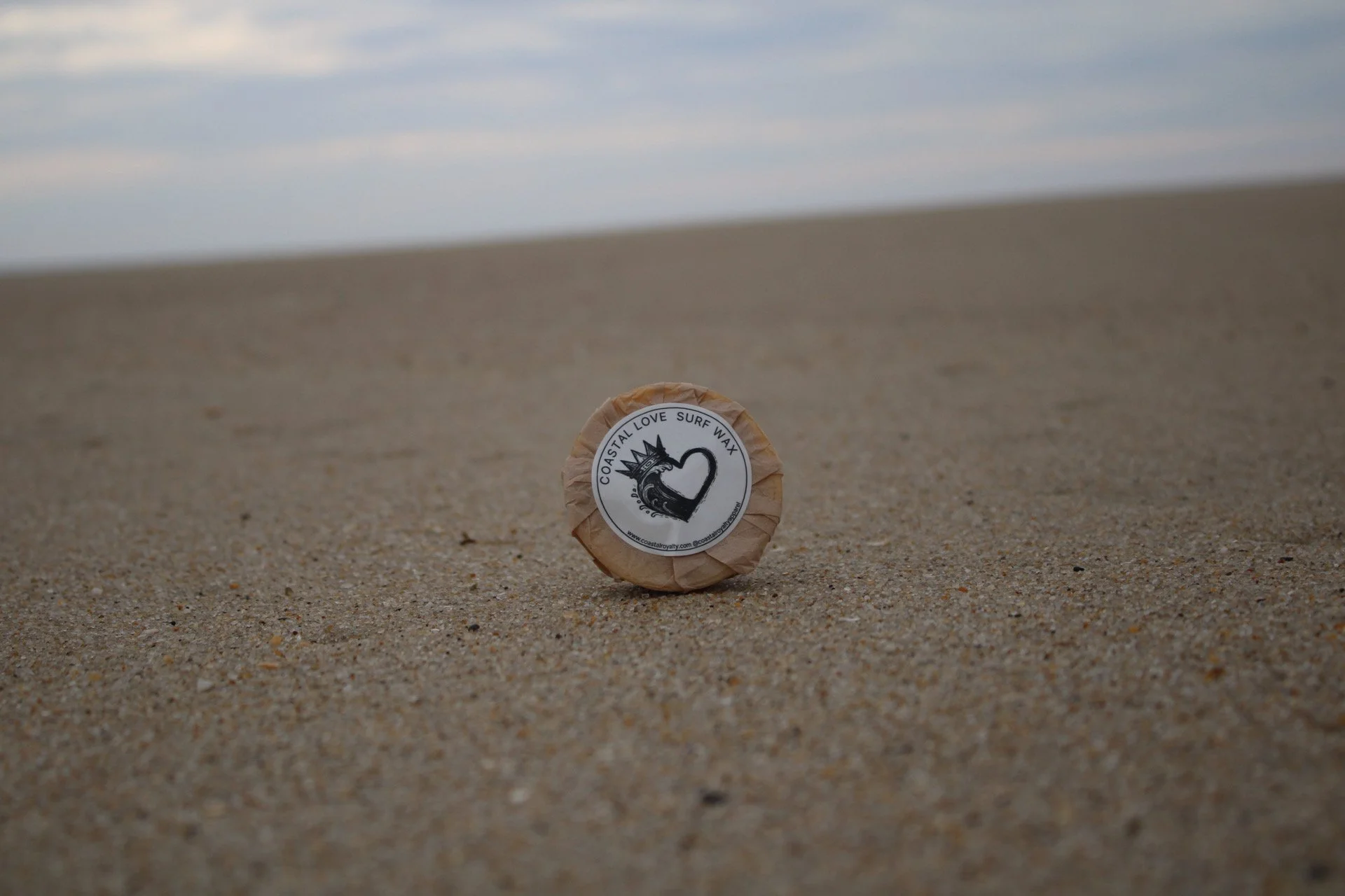 Coastal Love Surf Wax with sand and sky backdrop.
