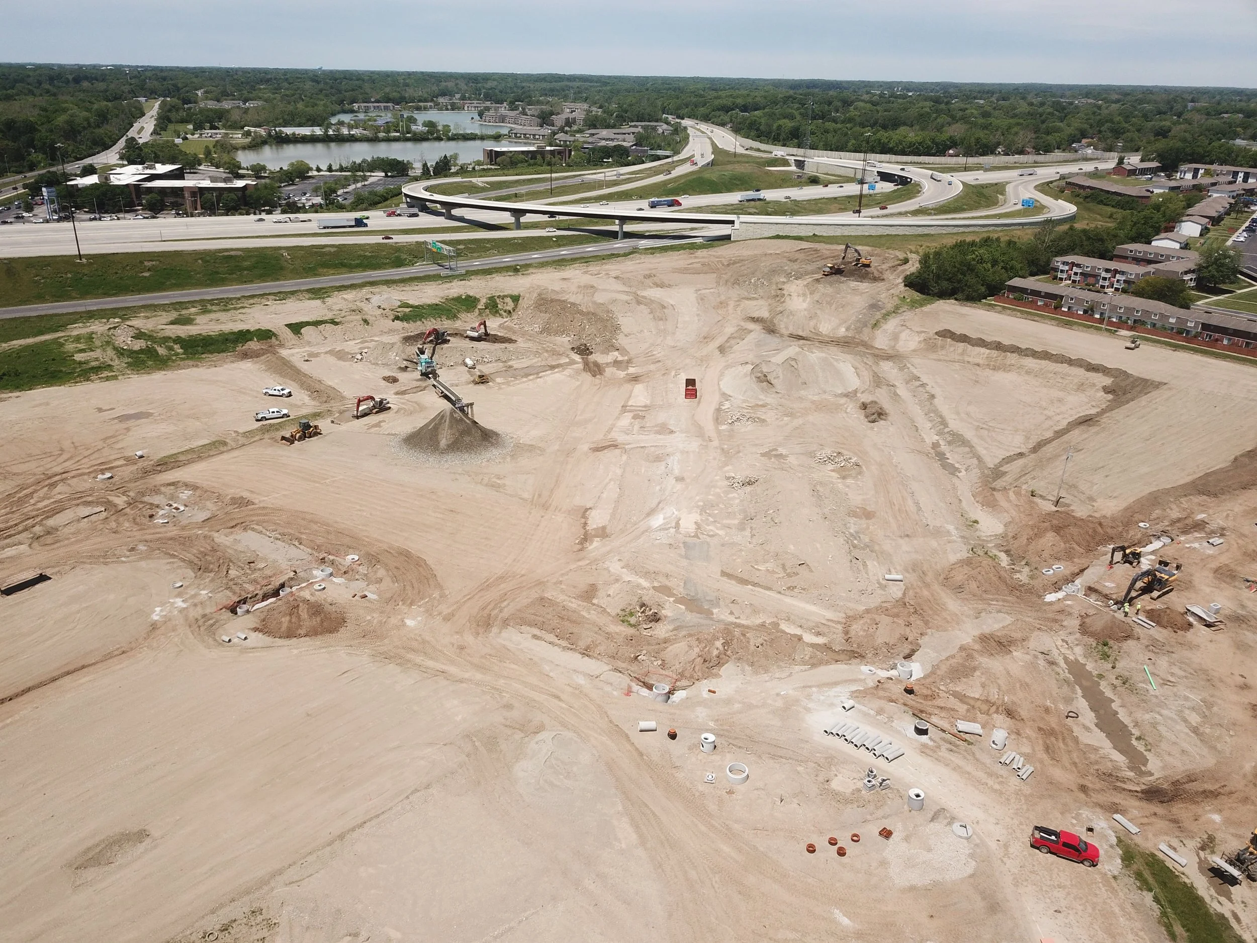 Aerial view of the Founders Square construction site before development with excavators, construction vehicles, and dirt piles, adjacent to a highway with vehicles and surrounding green landscape.