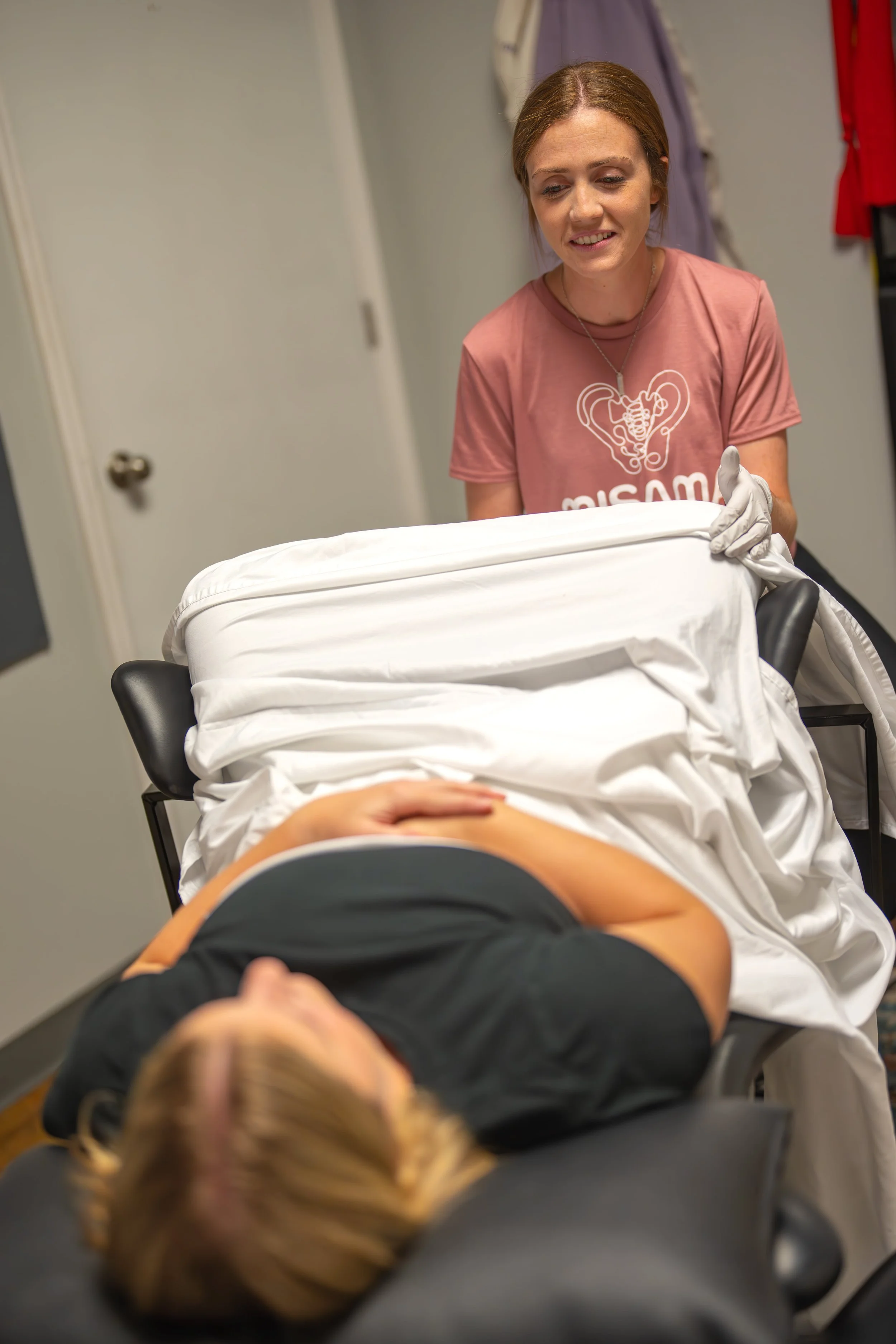 A woman lying on a hospital bed with her hands on her stomach, while another woman standing next to her adjusts the covers and looks down, inside a medical setting.