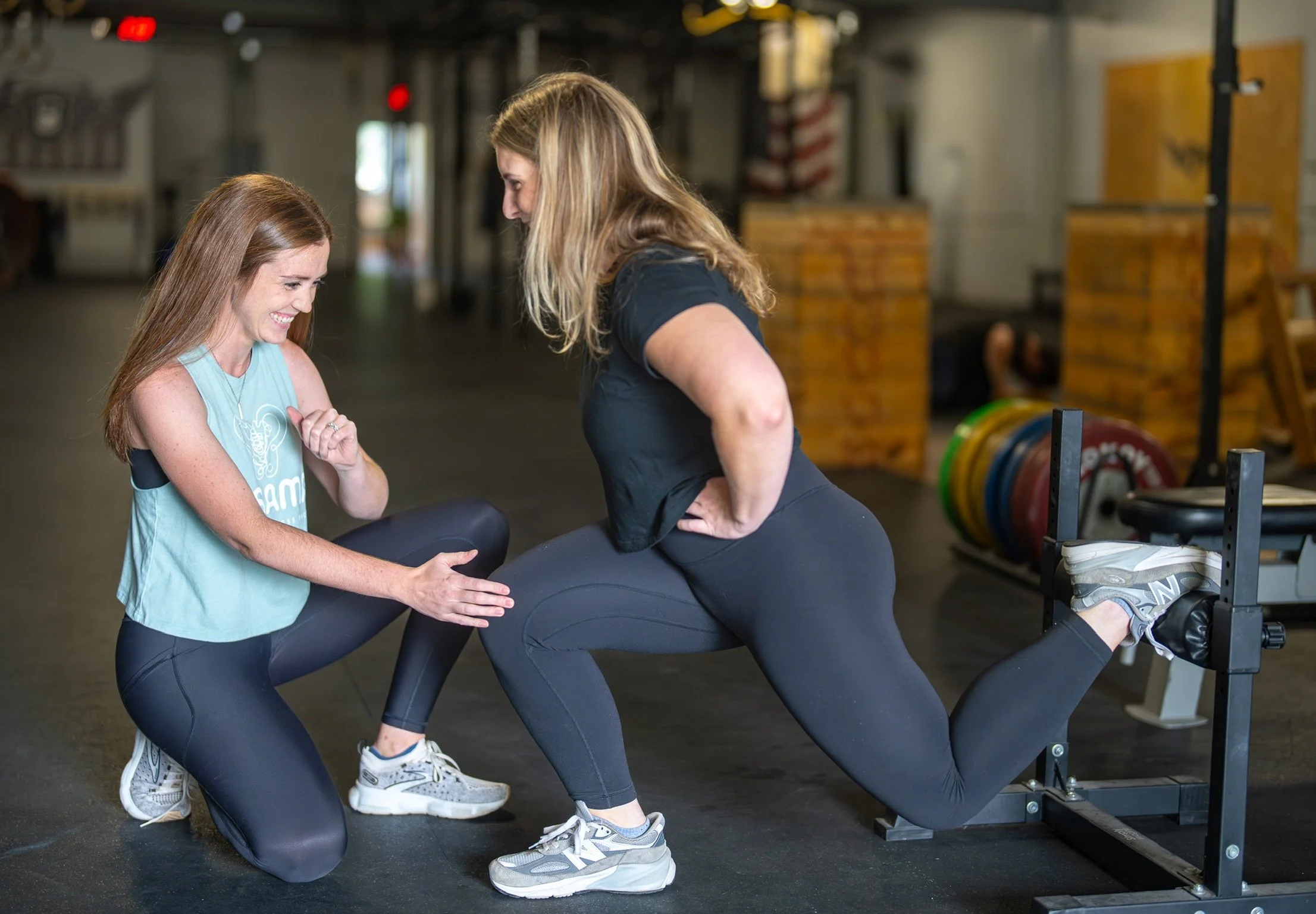 A woman in black leggings and sneakers is doing a kneeling exercise on a fitness machine, while a trainer in workout clothes is kneeling beside her, smiling, and providing encouragement in a gym setting.