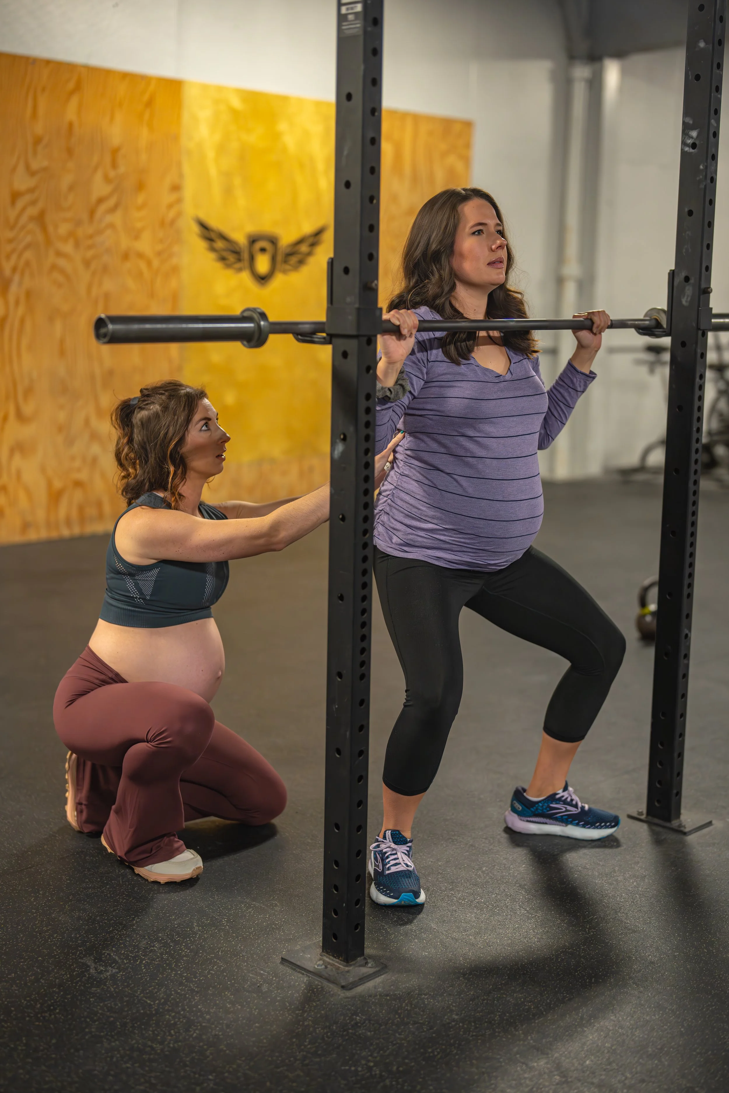 A woman in workout clothes performs a squat with a barbell in a gym, assisted by a trainer kneeling beside her.
