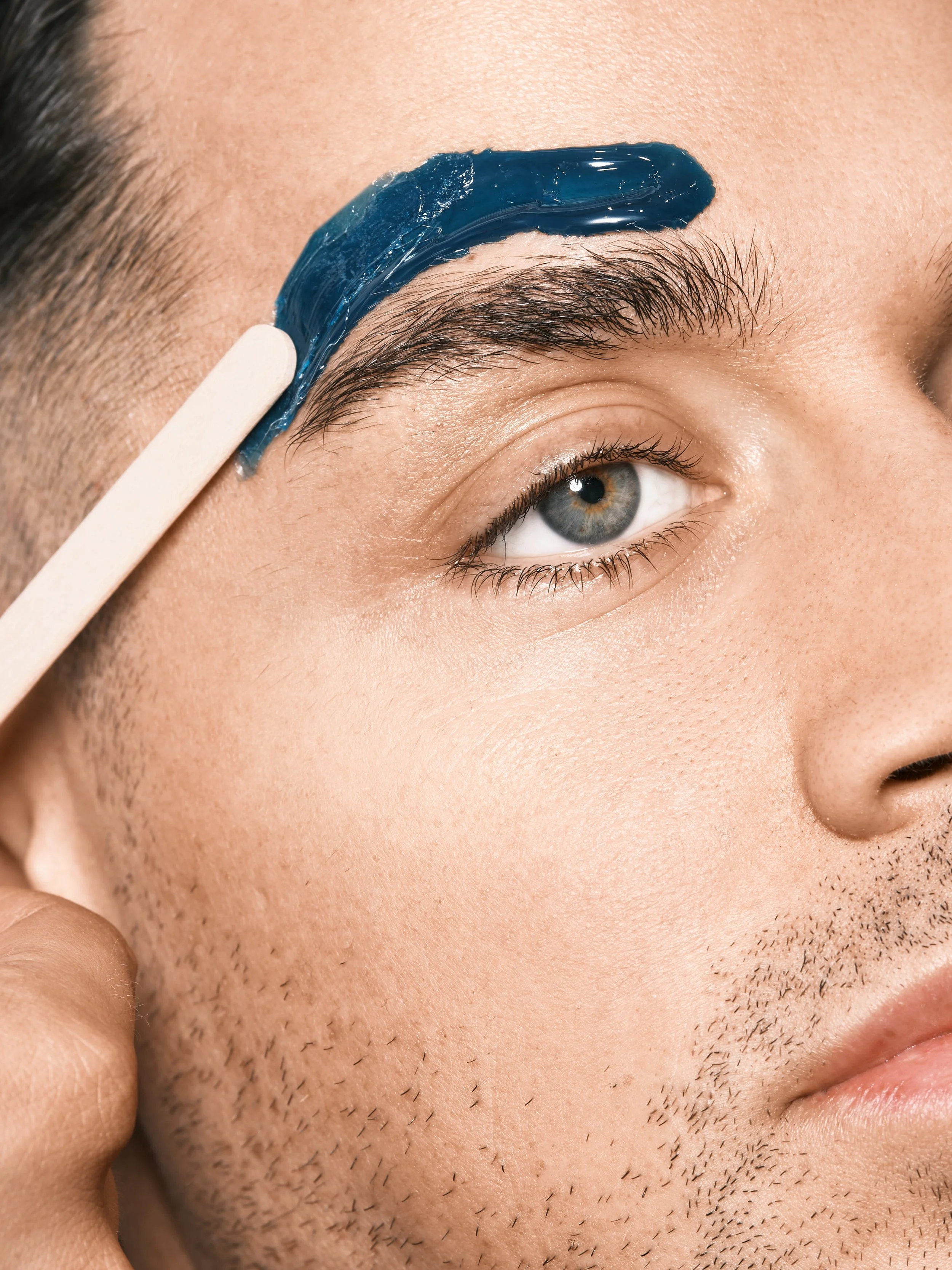 A close-up of a man's face as he applies blue facial mask to his eyebrow using a wooden spatula.