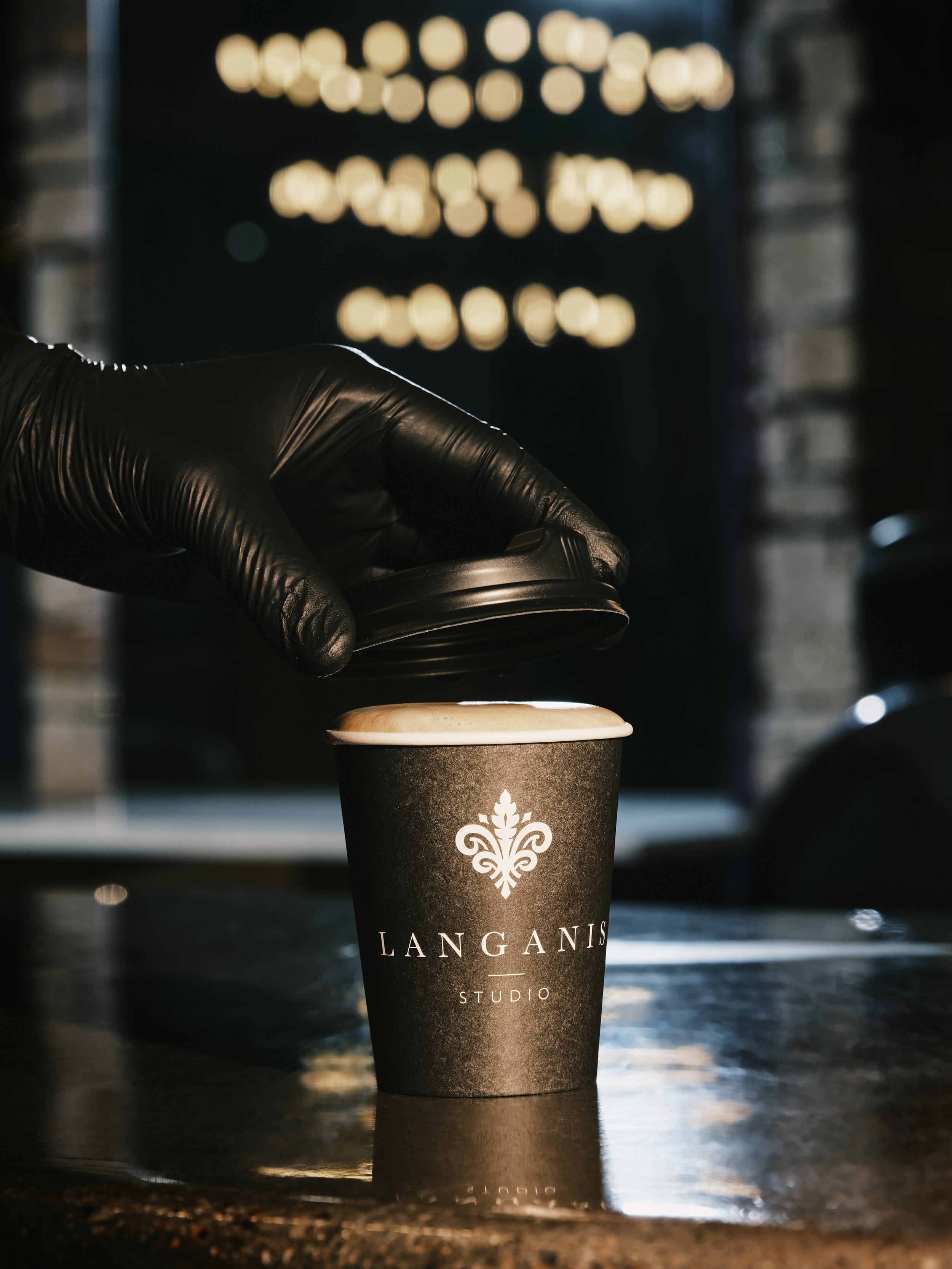A person wearing a black glove opening a black paper coffee cup with white foam at the top, sitting on a dark reflective surface at a coffee shop. The cup has a logo with the text 'Langan's Studio'.