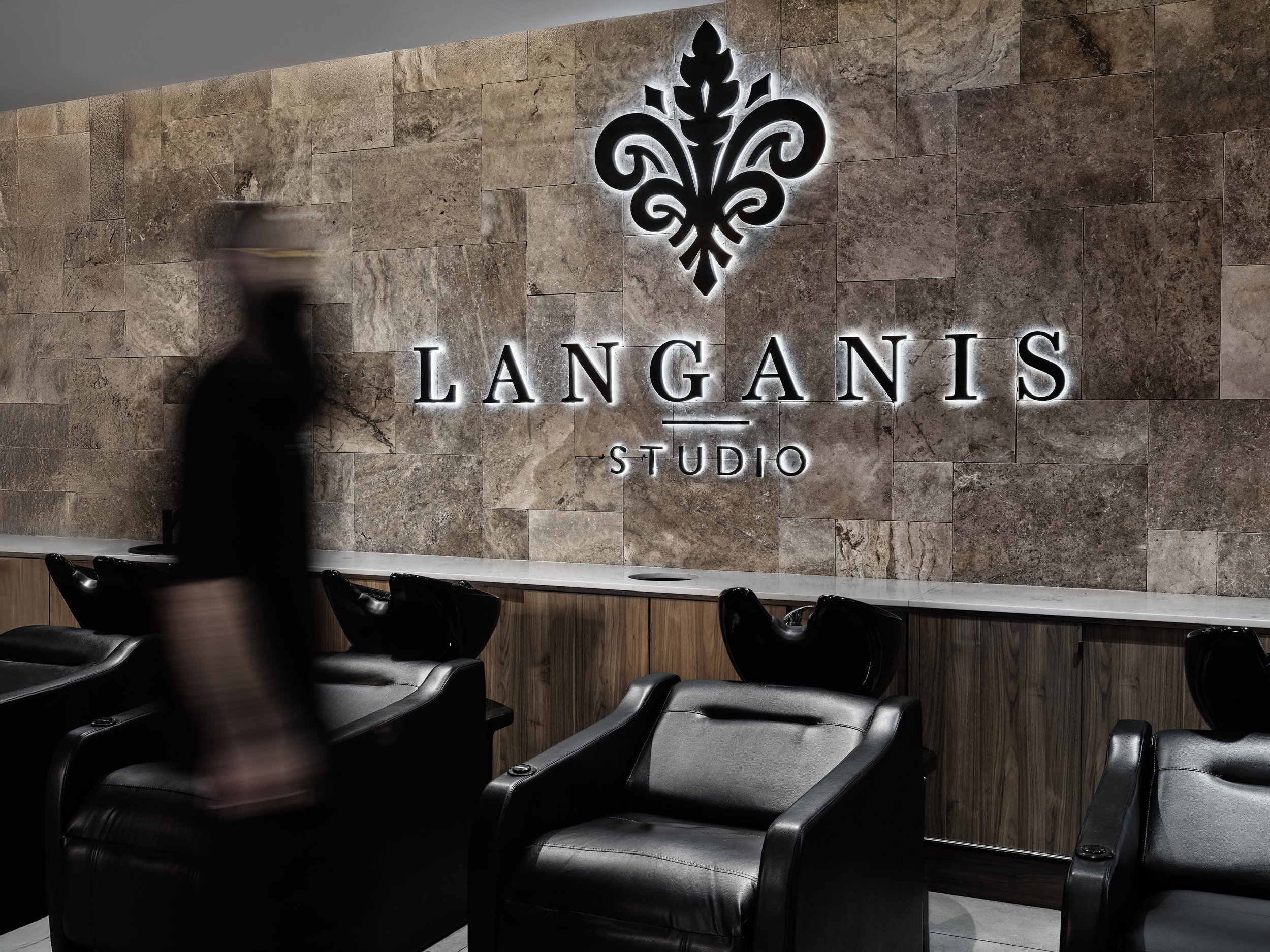 Interior of a salon or spa with black chairs in front of a marble wall, illuminated sign reads 'LANGANIS STUDIO' with a decorative logo above.