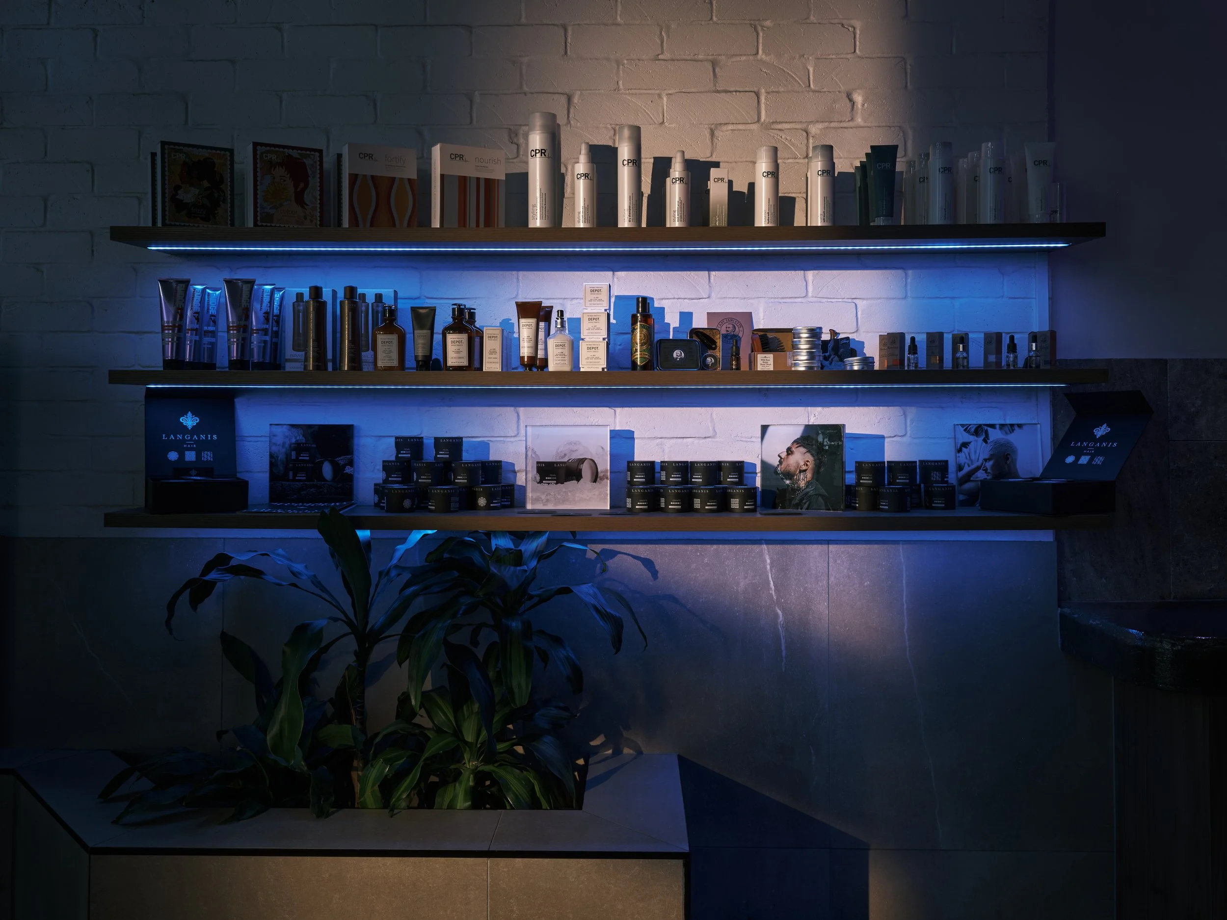 Display of hair and skincare products on illuminated shelves against a white brick wall with a potted plant in the foreground.