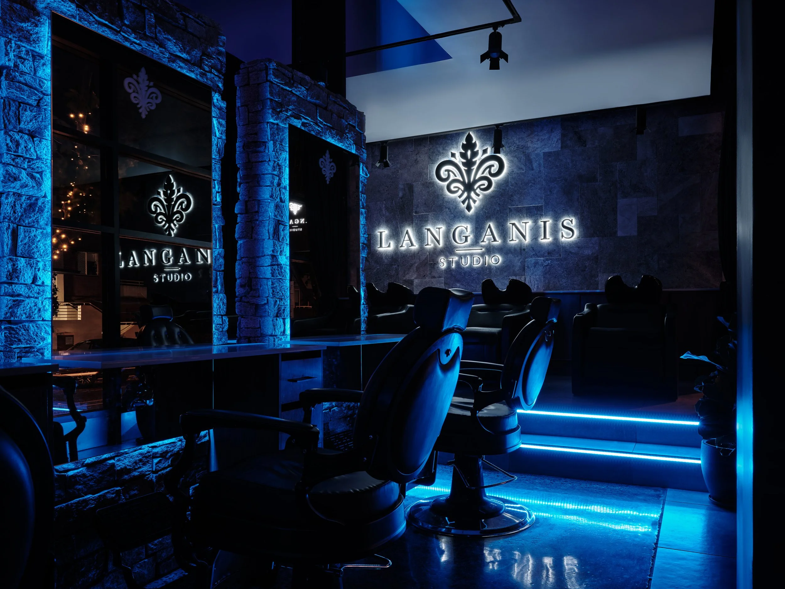 Interior of a modern studio with black chairs, a large illuminated sign reading 'LANGANIS STUDIO' on a textured wall, and blue accent lighting.