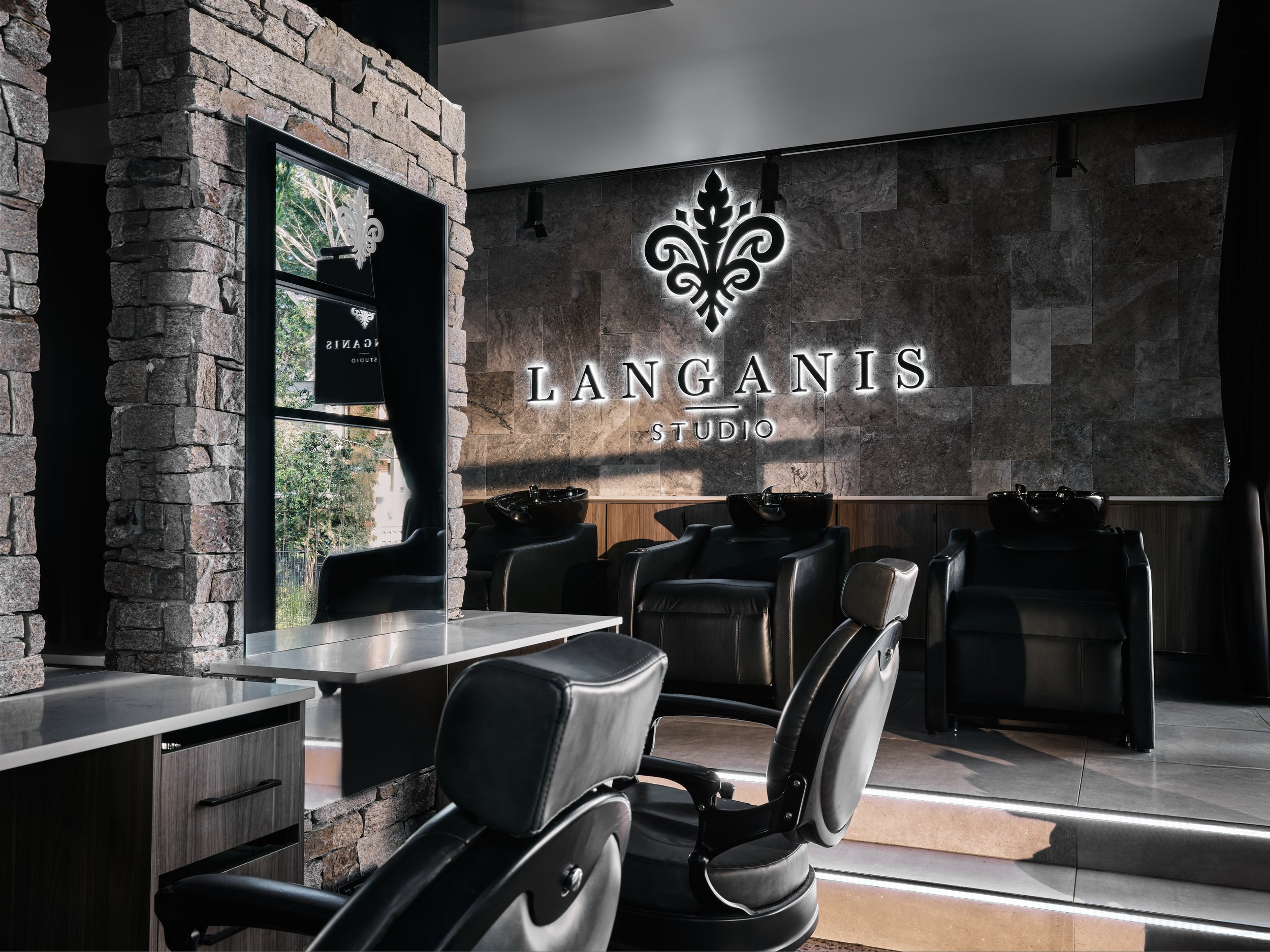 Interior of a beauty salon or spa with black hair washing stations, a large mirror, and a stone accent wall featuring a illuminated sign that reads 'LANGANIS STUDIO' with a decorative emblem.