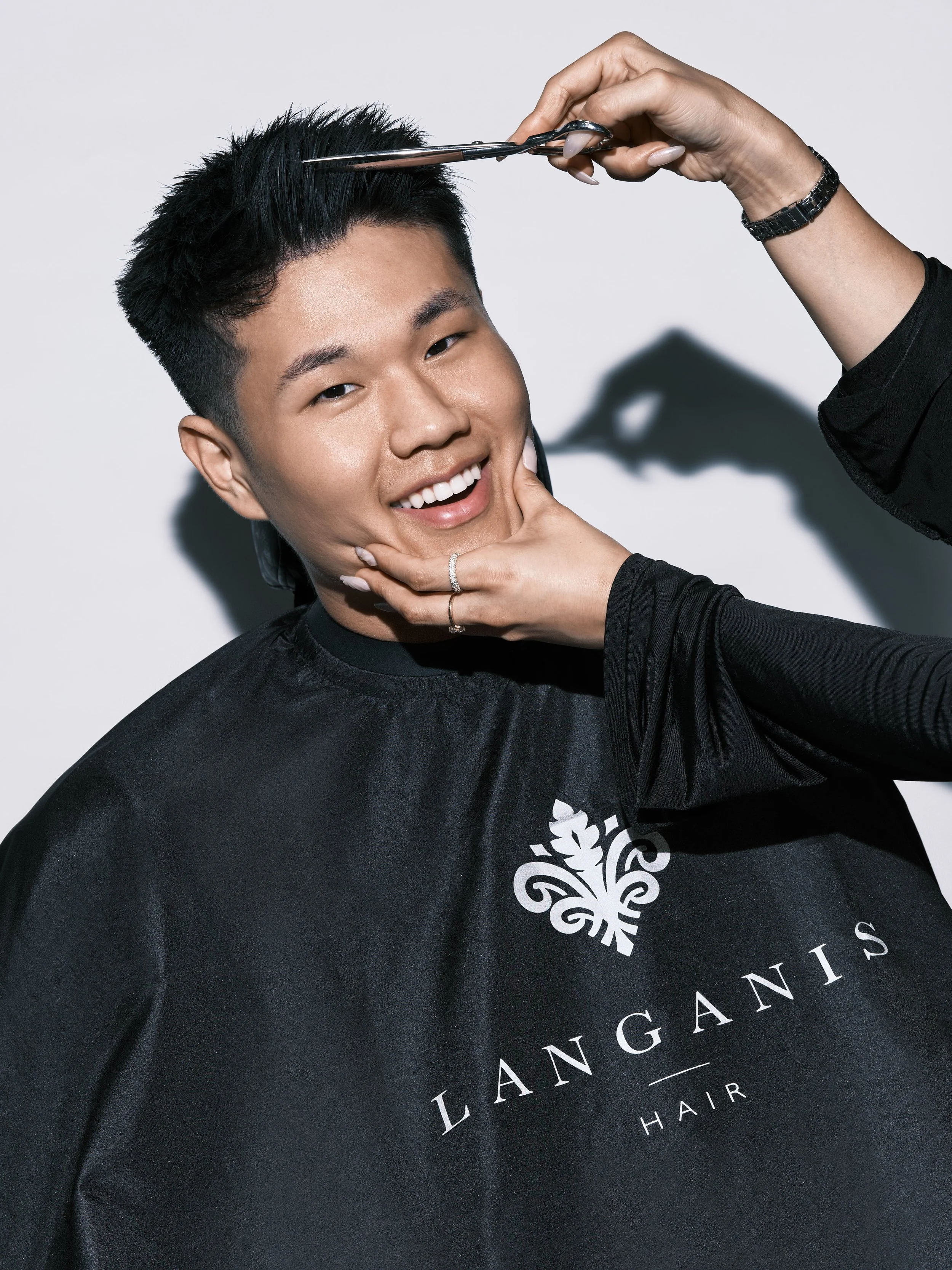 A person with short, black hair smiling during a haircut. A stylist's hand is visible, holding scissors to trim the hair. The person is wearing a black salon cape with a white logo that reads 'LANGANIS HAIR'.