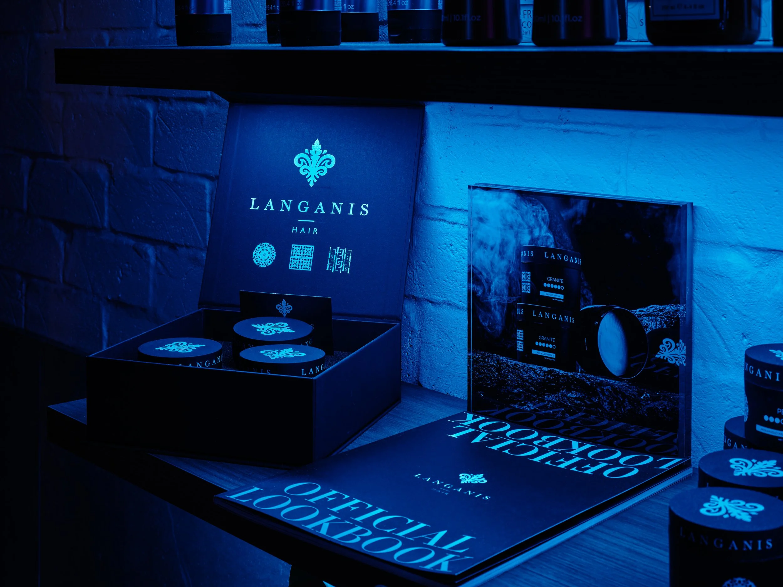 Display of Langanis hair products with black packaging, placed on a wooden surface, illuminated by blue lighting, with a brochure and a mirror reflecting the product packaging, against a blue brick wall.