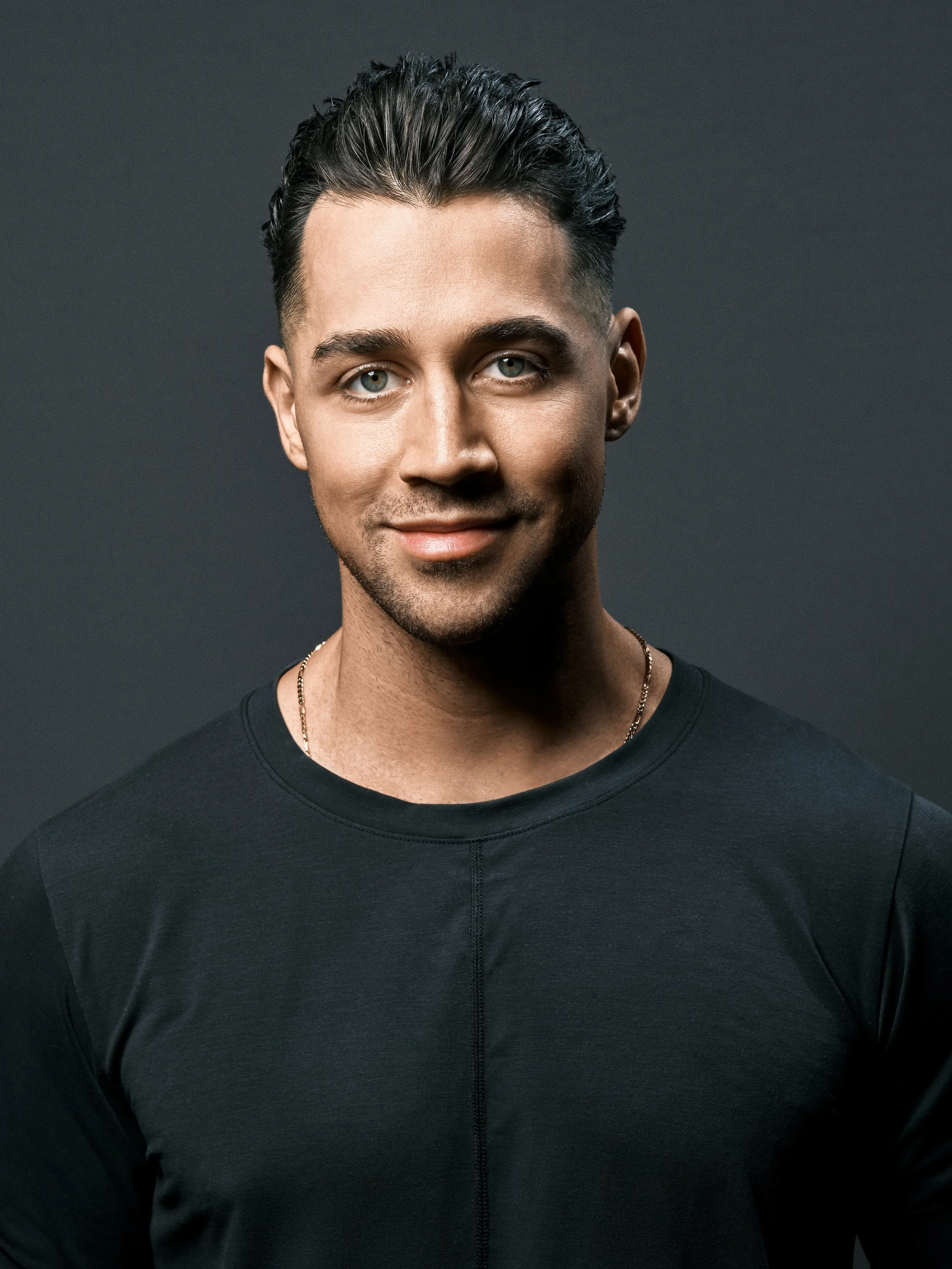 Portrait of a young man with dark hair and light eyes, wearing a black shirt against a dark background.