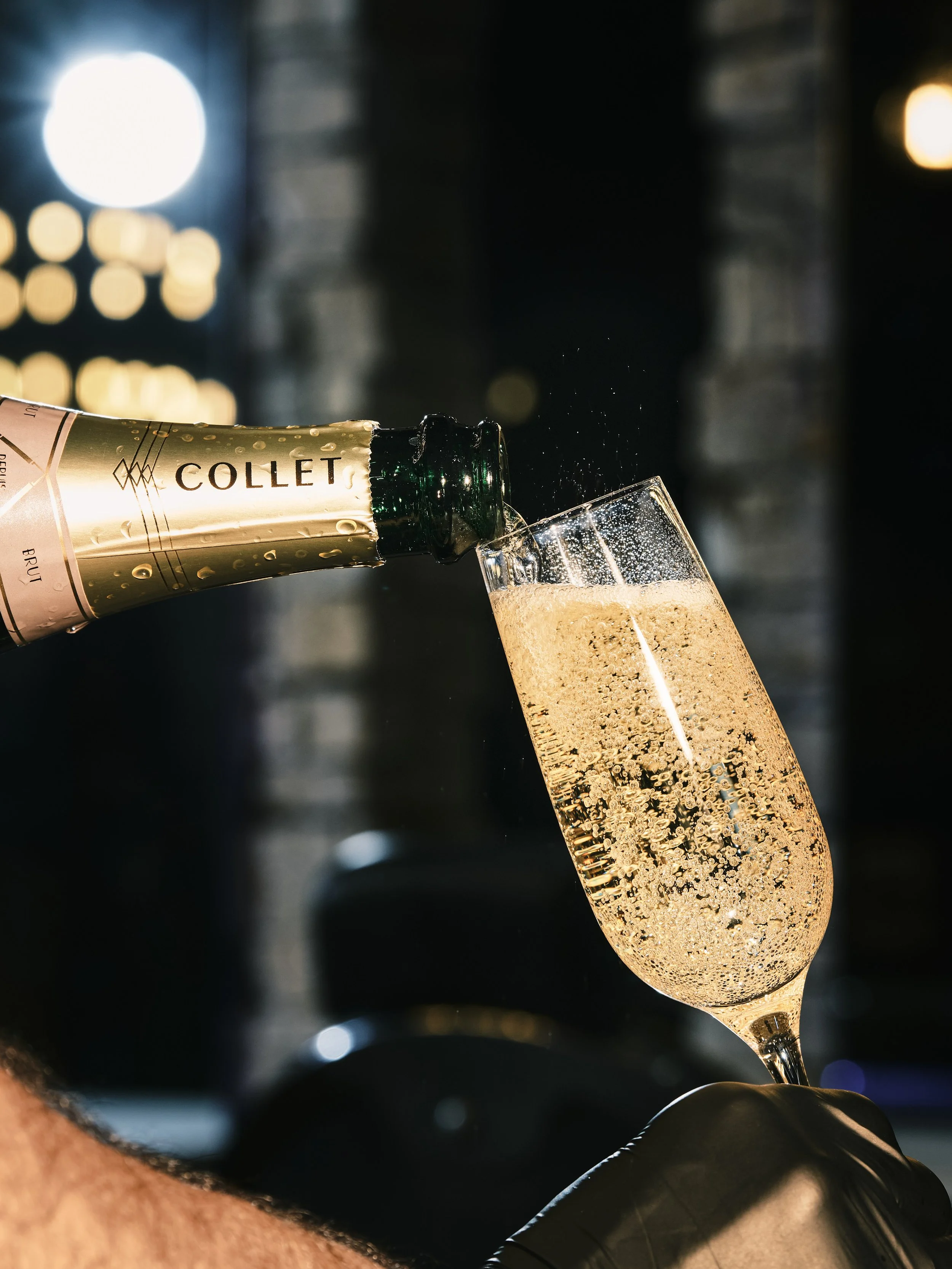A hand holding a champagne glass while champagne is being poured from a bottle labeled 'COLLET'. The background is blurred with warm lights and dark surroundings.