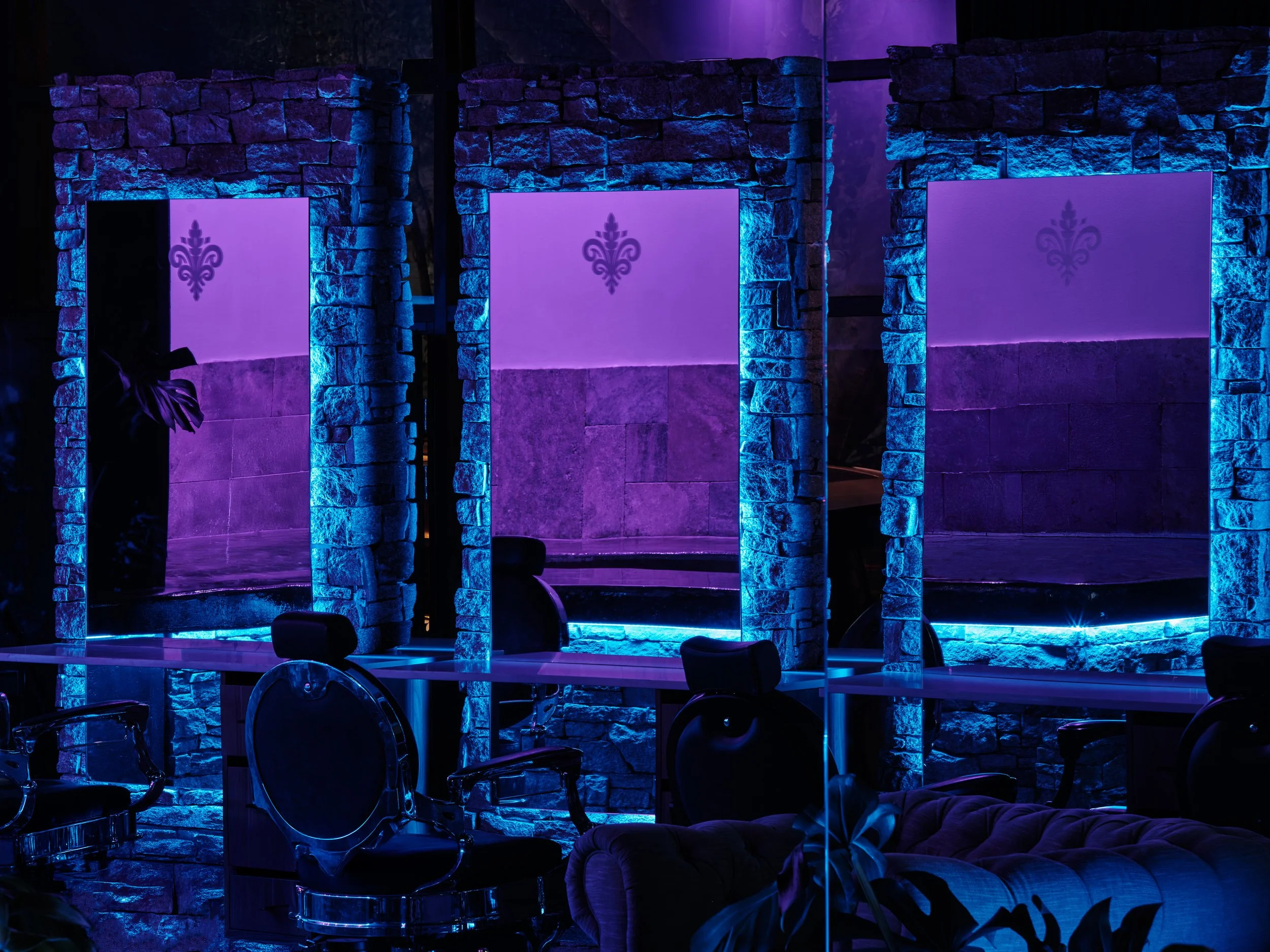 Three illuminated dressing room mirrors with blue lighting, stone walls, black chairs, and a purple flower decoration.