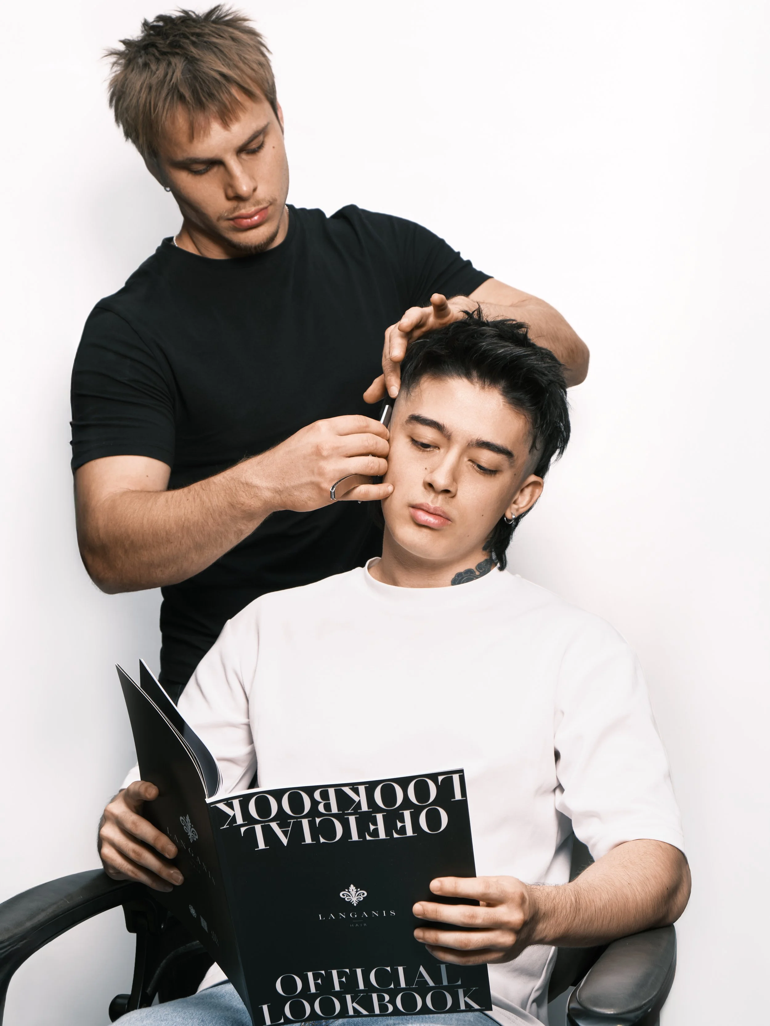 A hairstylist is cutting a young man's hair while he reads a magazine titled 'Official Lookbook' with the logo 'Langanis'.