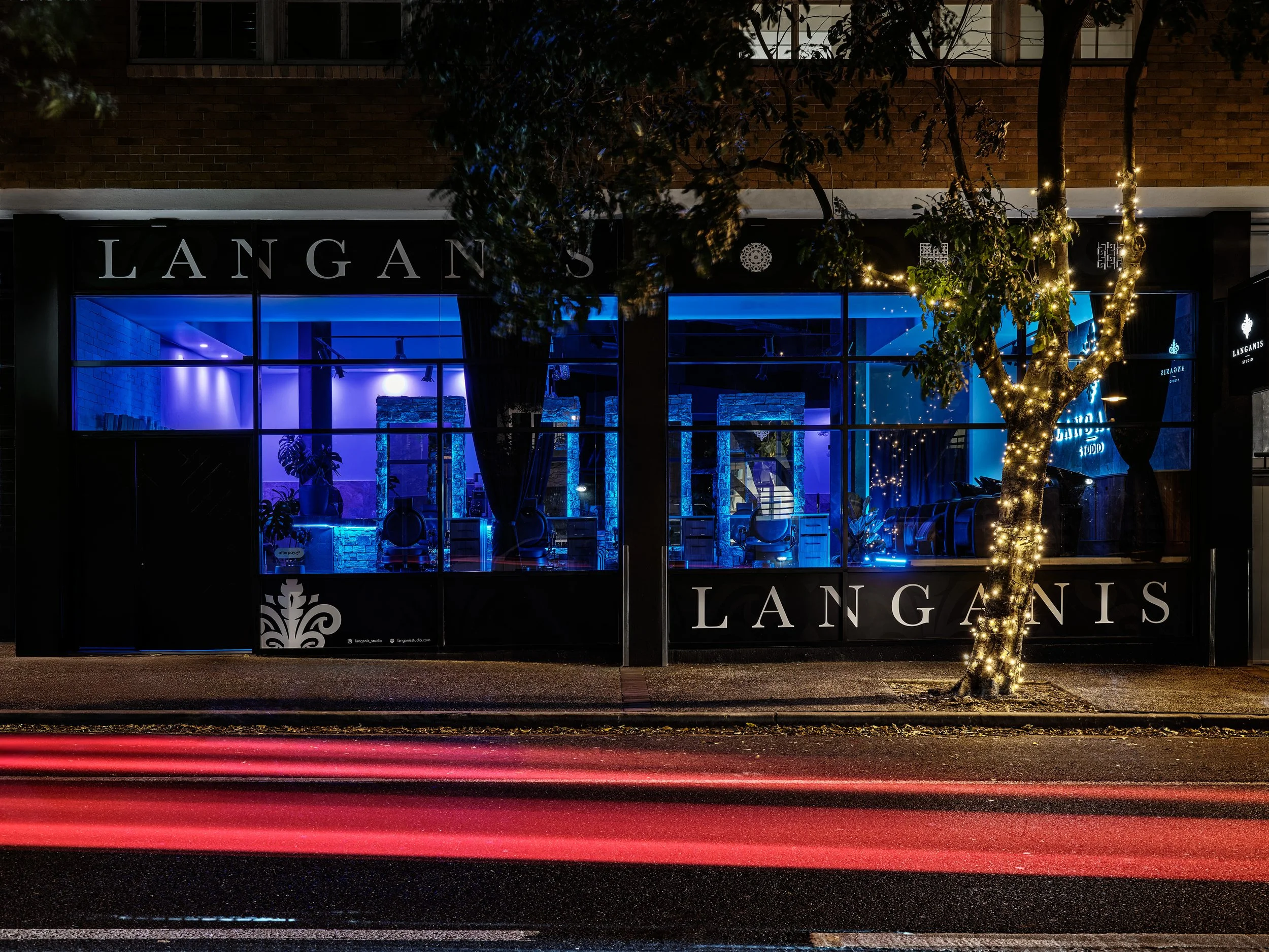 Nighttime view of a storefront with large glass windows illuminated with blue lighting, and a tree wrapped in string lights in front. The store name is 'LANGANIS' displayed on the window.