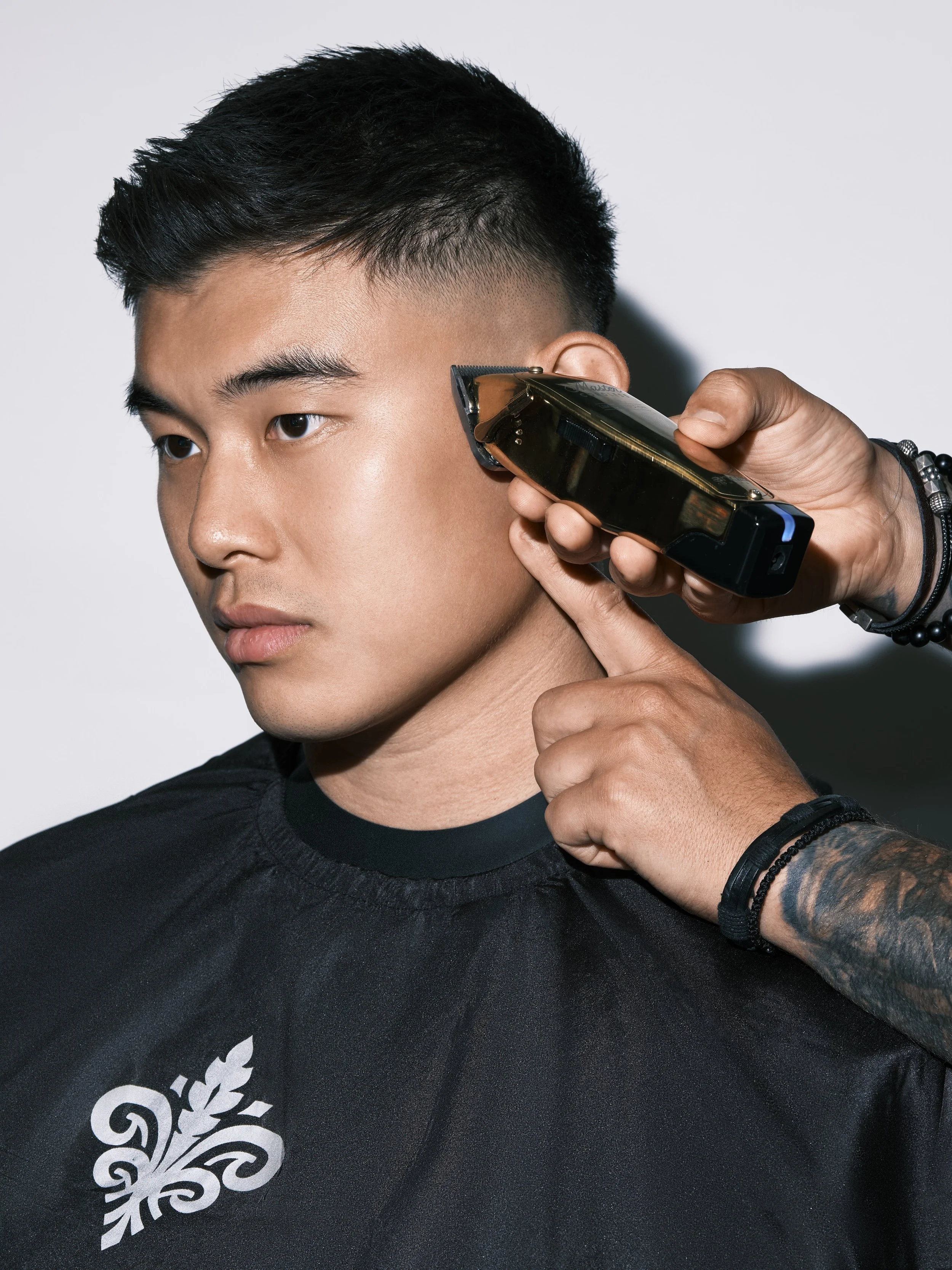 A man getting a haircut with electric hair clippers, wearing a black cape with a white logo, against a plain background.