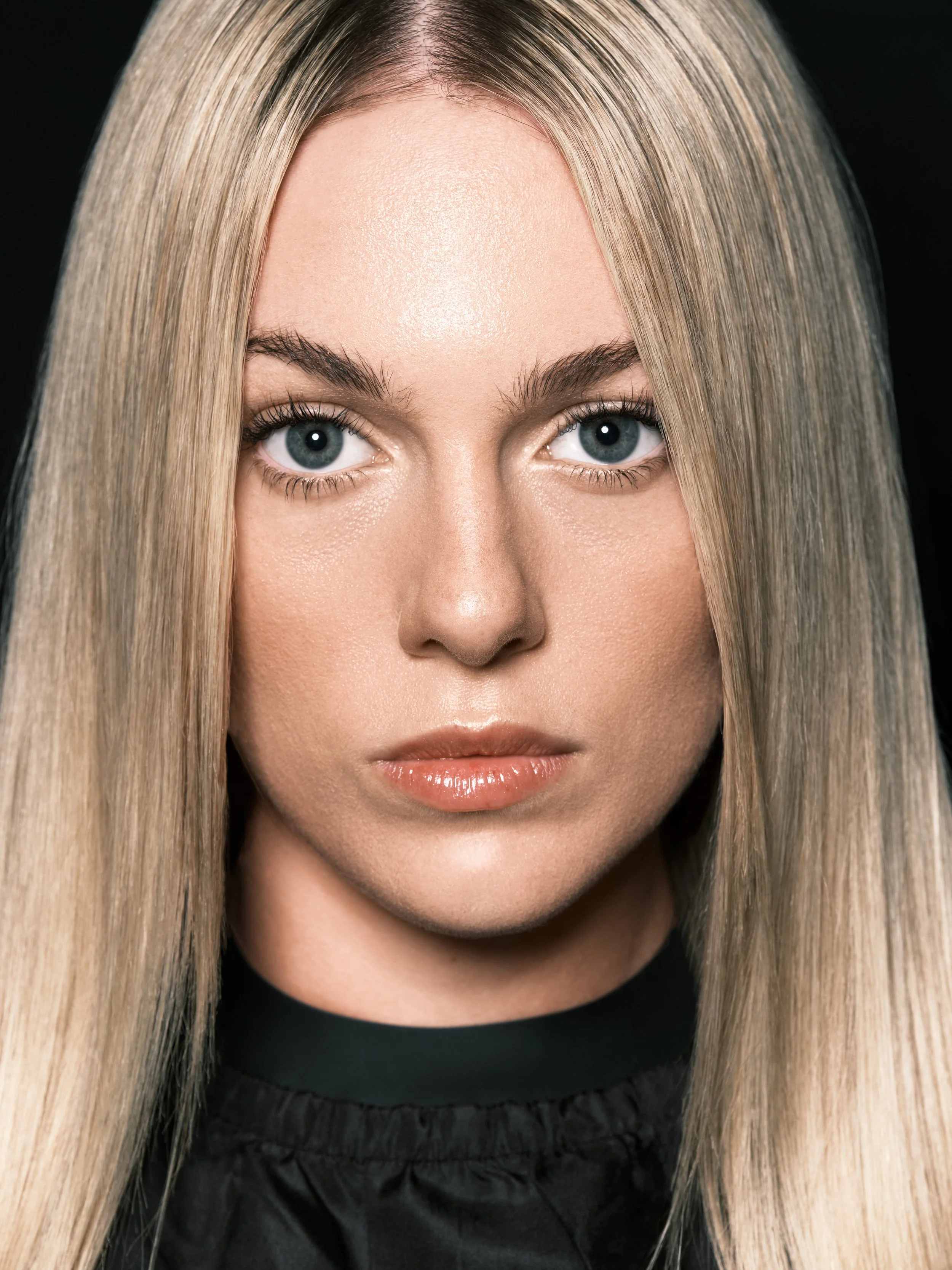 Close-up portrait of a young woman with long blonde hair, blue eyes, and light makeup, looking directly at the camera against a dark background.