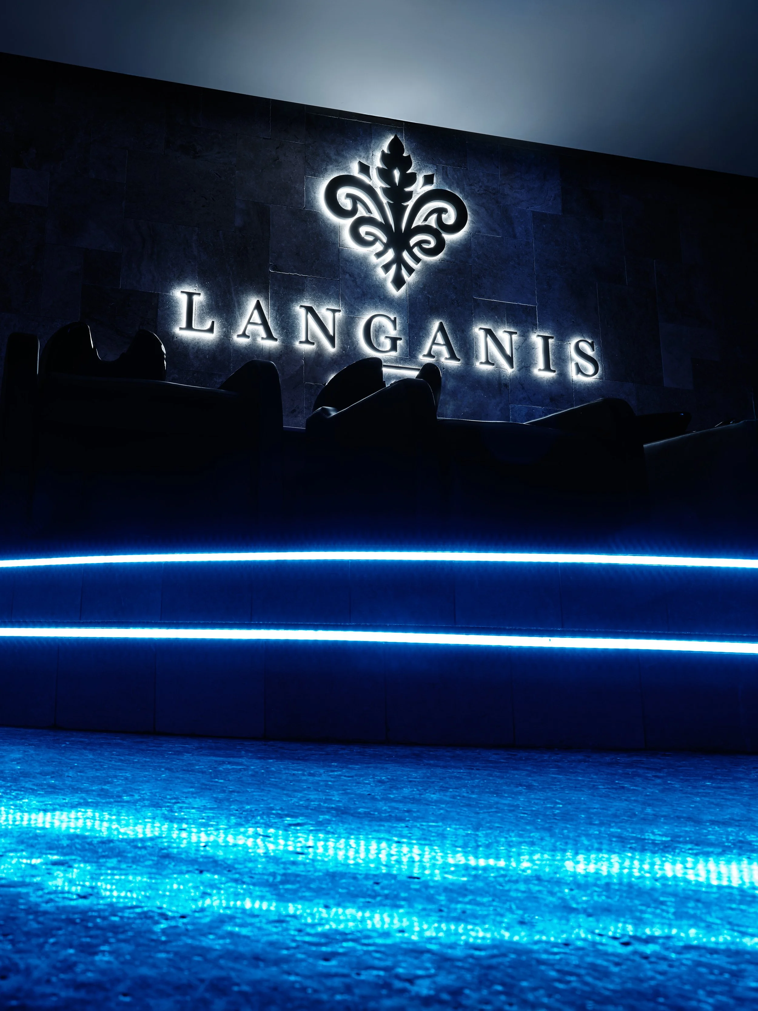 Neon-lit sign reading 'LANGANIS' with a decorative emblem above, on a dark wall, with dim seating and blue accent lighting.