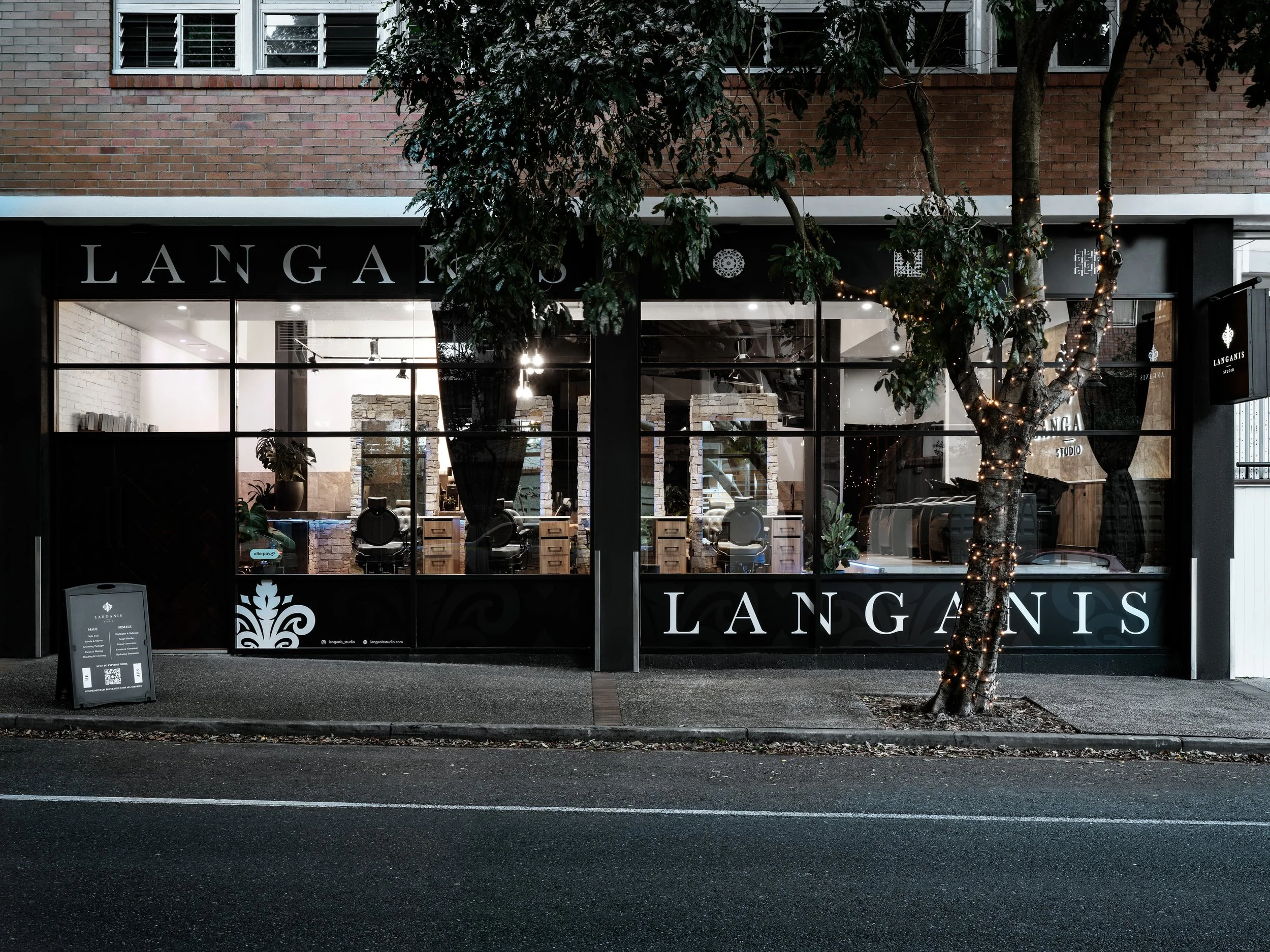 Storefront with large glass windows displaying interior furniture and plants, with the name 'LANGAINS' on the window and the storefront sign. A tree with string lights is outside, and a sidewalk sign is on the sidewalk in front of the store.