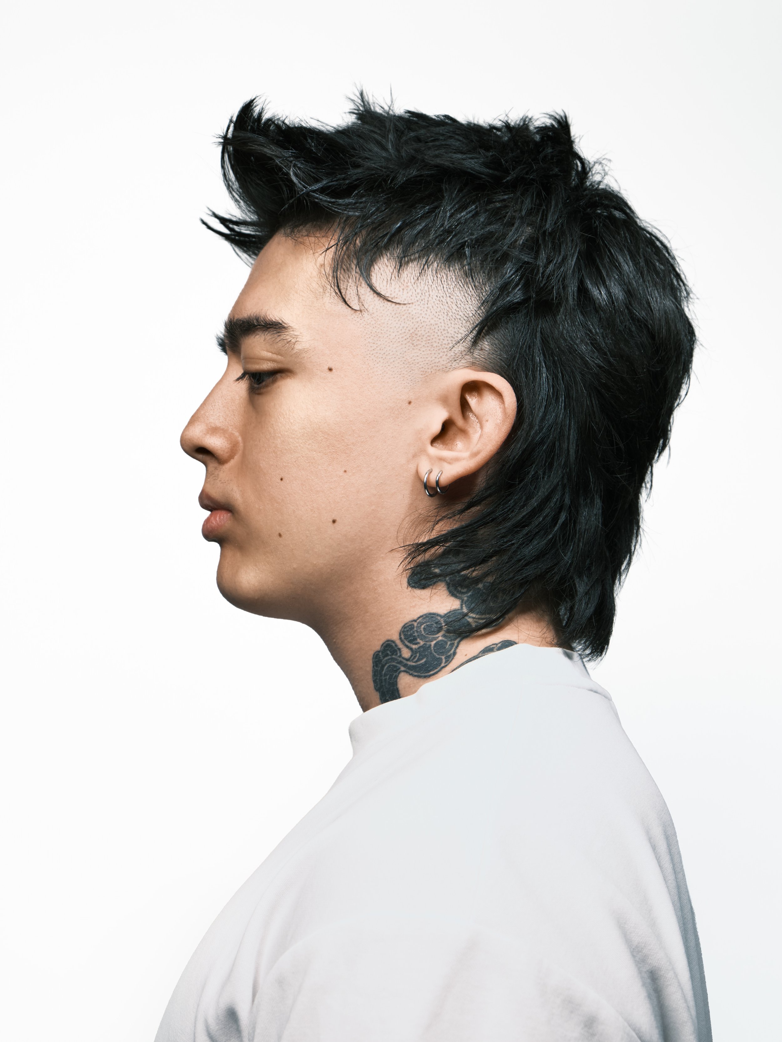 Side profile of a person with black, short, messy hair and multiple earrings, wearing a white shirt, showing a tattoo of clouds or smoke on their neck, against a plain white background.