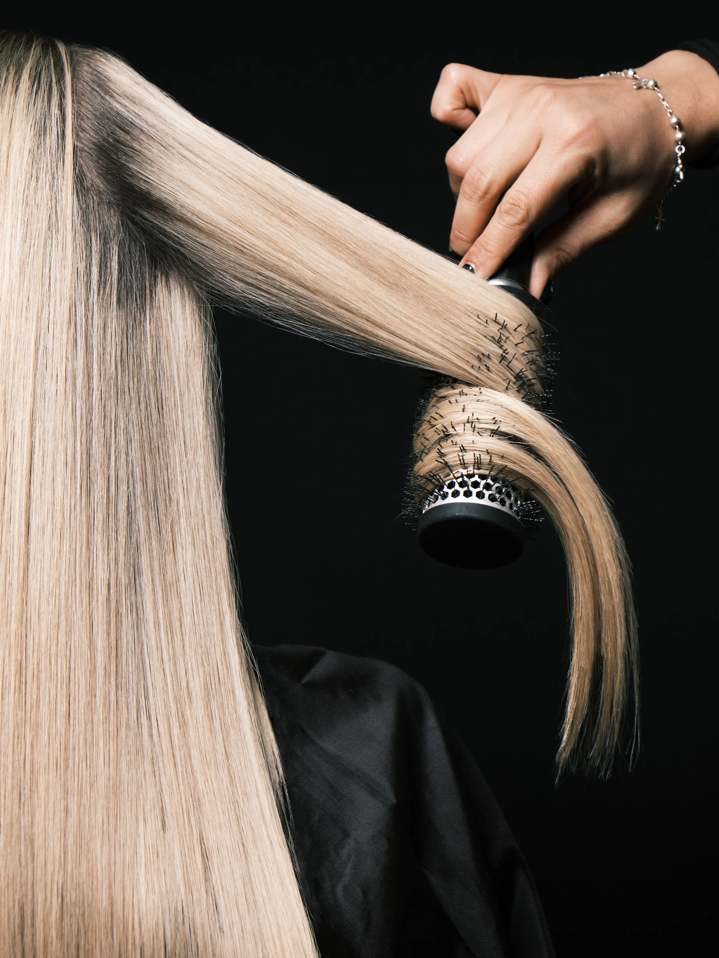 A person with blonde hair is having their hair blow-dried with a round brush and a hairdryer, against a dark background.