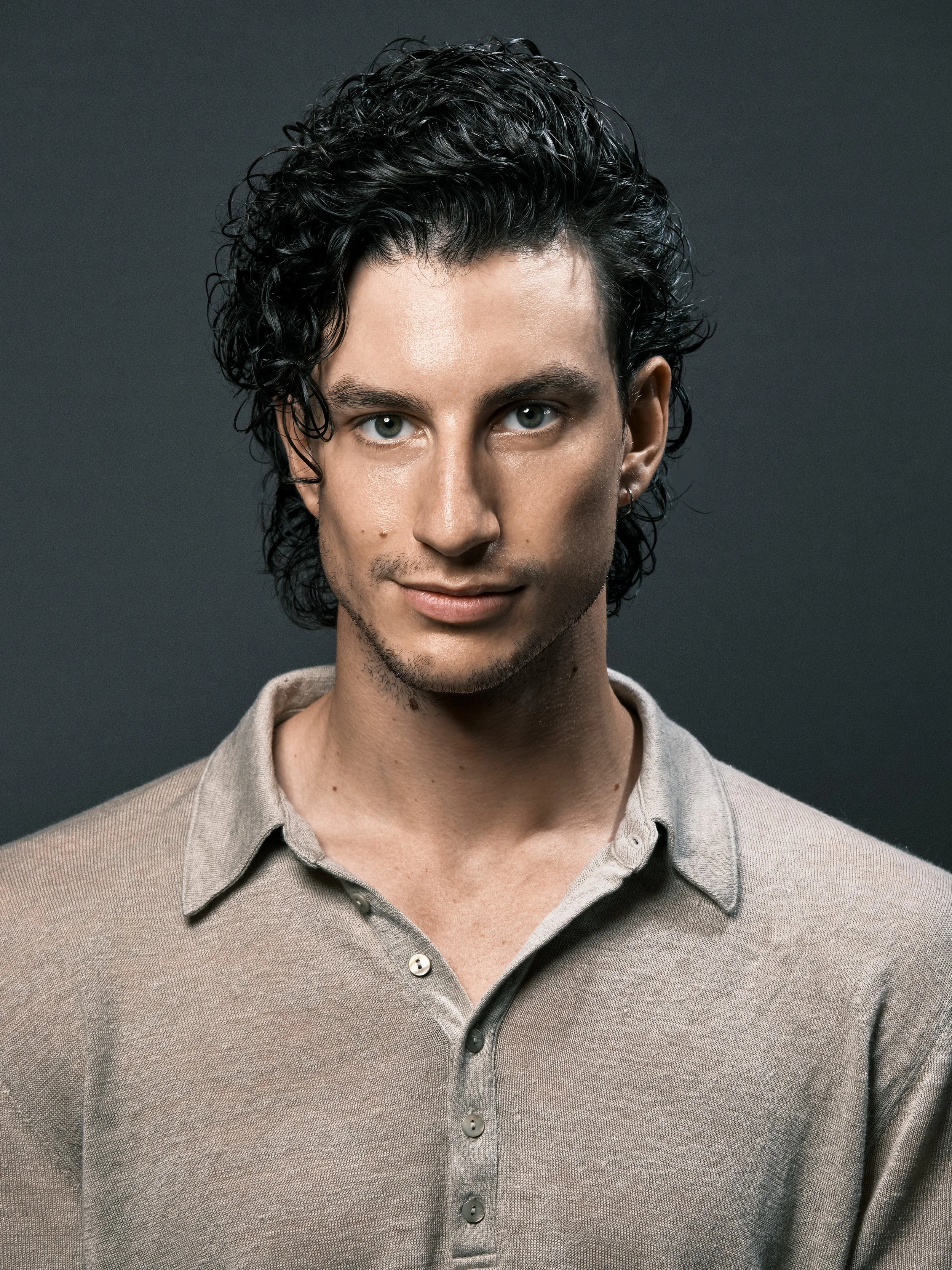 A young man with curly black hair, blue eyes, and light skin, wearing a beige button-up shirt, looking at the camera against a dark gray background.