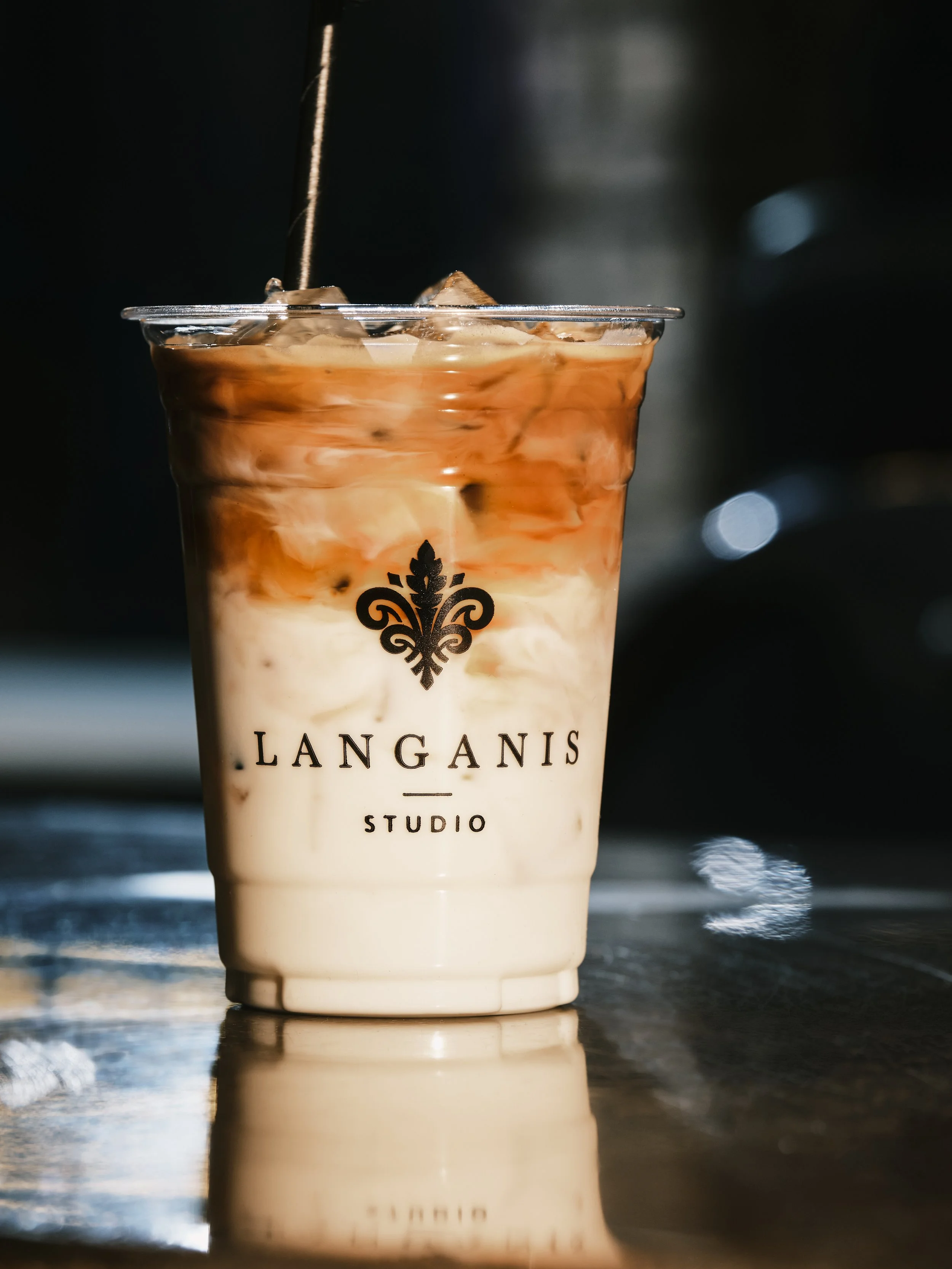 Iced coffee with milk and chocolate syrup in a clear plastic cup labeled 'LANGANIS STUDIO' with ice cubes on a reflective surface.