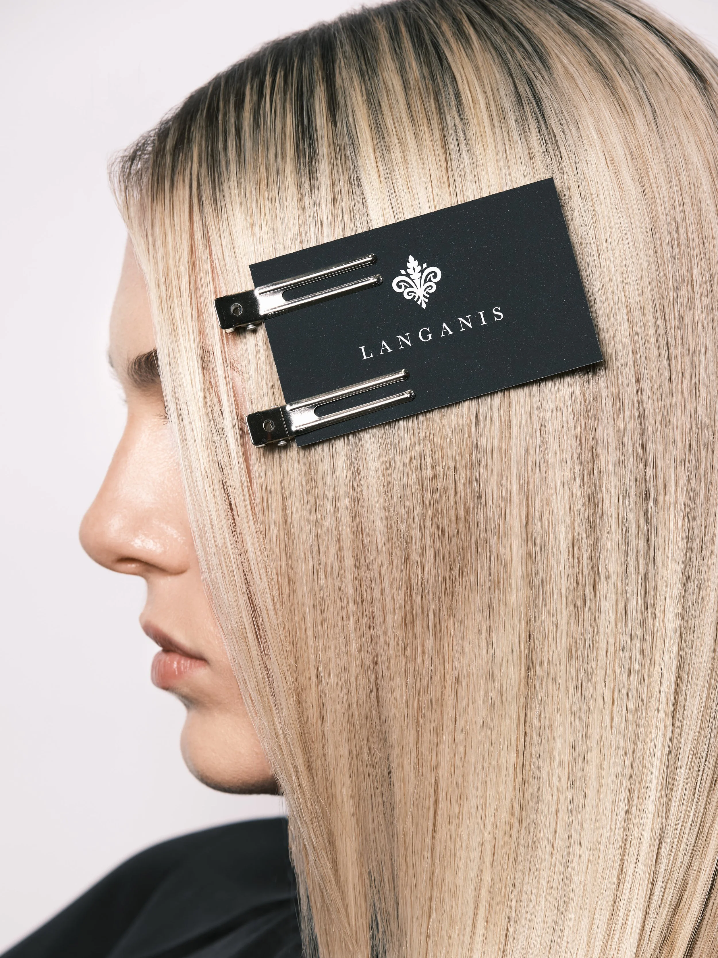 Side profile of a woman with sleek, long blonde hair secured with a black hair clip bearing a small black card with the words "LANGANIS" and a decorative emblem.
