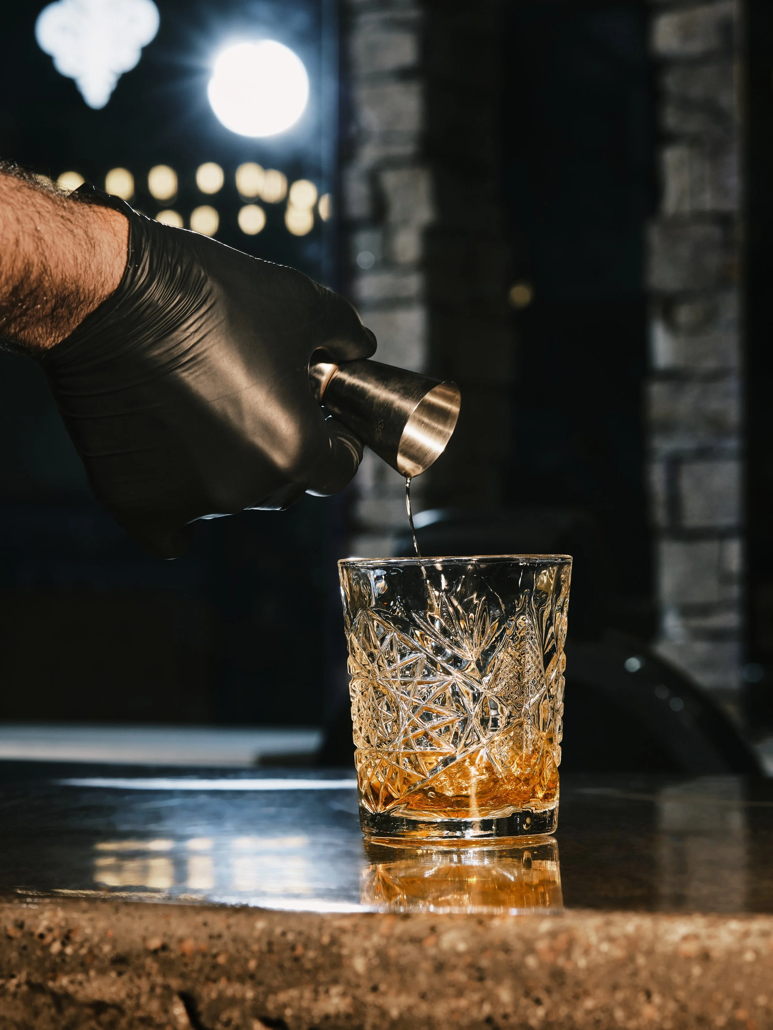 A person wearing a black glove pouring liquid from a jigger into a glass with a textured pattern on a bar counter, with a dark and brick background.
