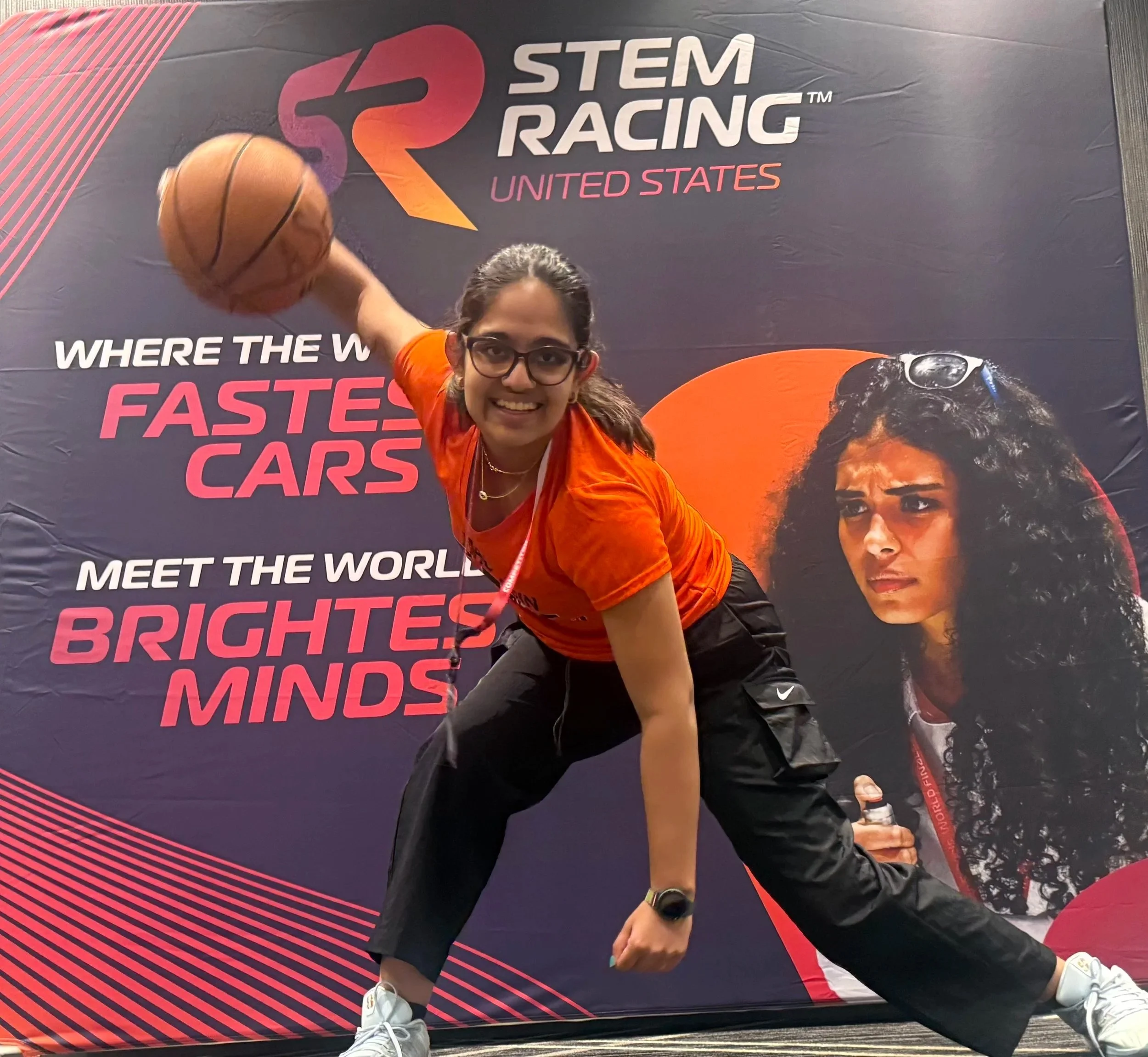 Ashna smiling indoors, wearing glasses, an orange shirt, and black athletic pants, handling a basketball in front of the STEM Racing backdrop.
