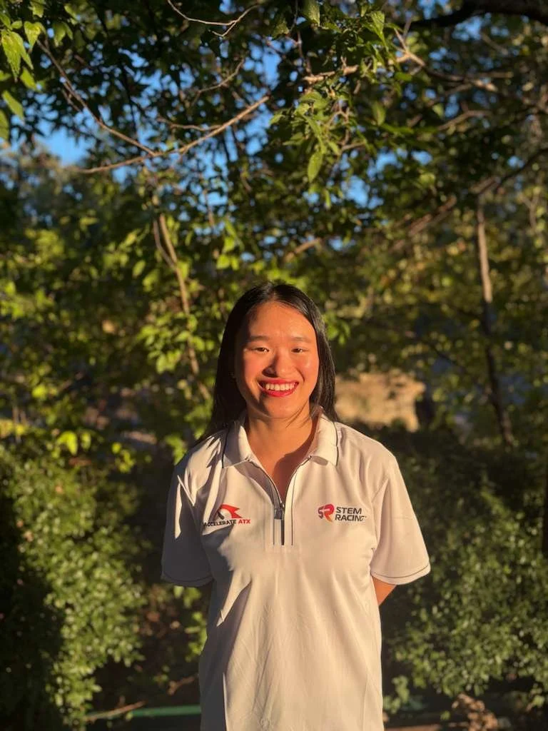 Kylie smiling outdoors, wearing a white sports polo with 'Accelerate ATX' and 'STEM Racing' logos, standing in front of green leafy trees during daylight.
