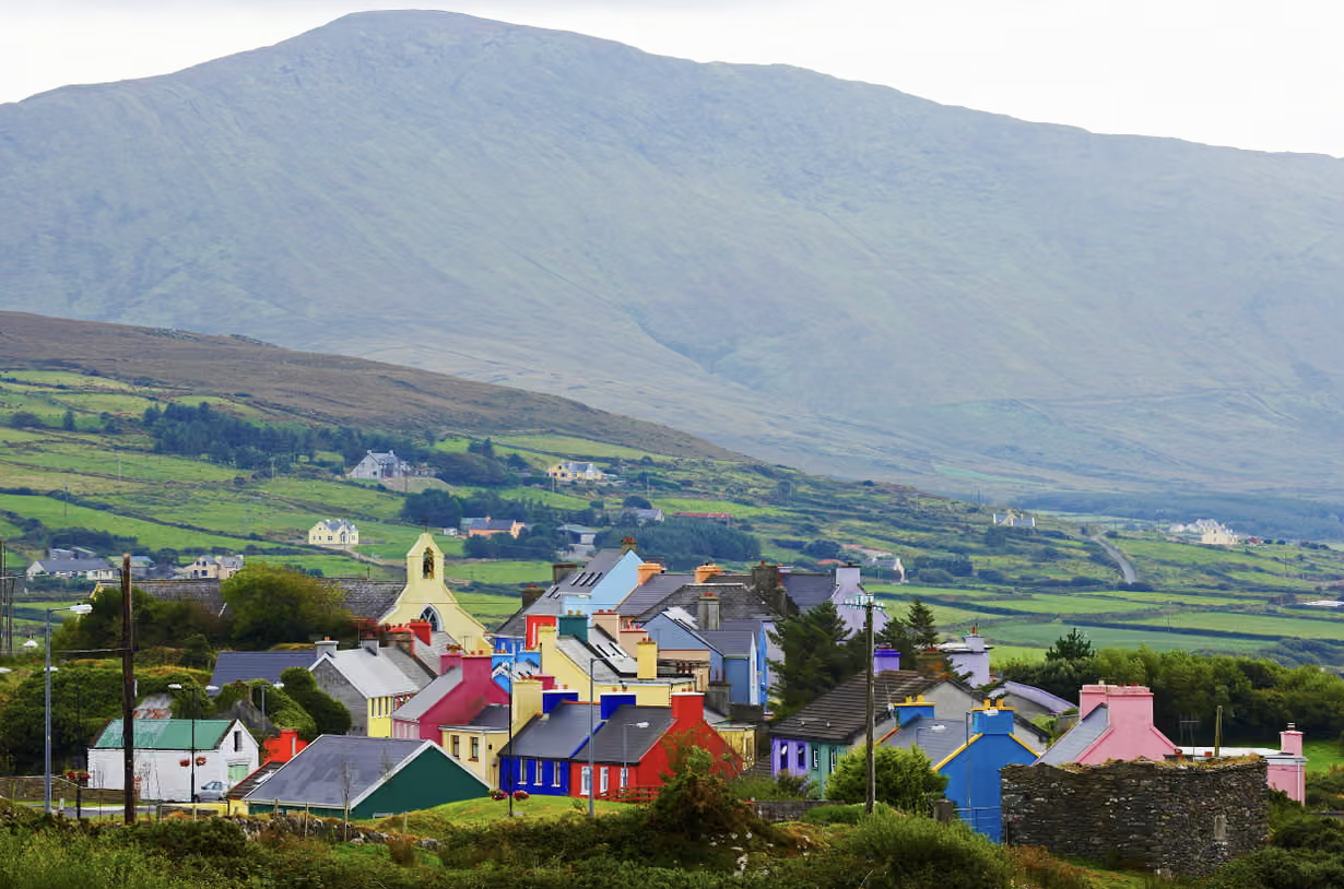 Colorful houses in a small village in south Ireland with a mountain in the background, green fields surrounding the village.