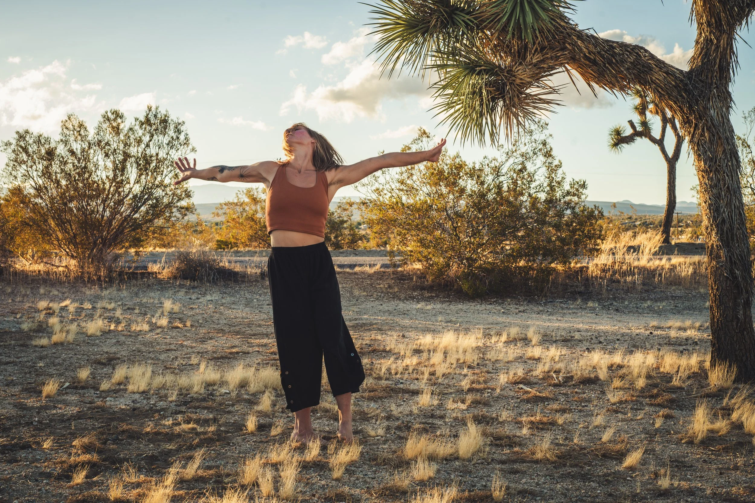 A woman standing in a desert landscape with her arms wide open and her face turned upwards, surrounded by dry bushes and a tree, during sunset or sunrise.