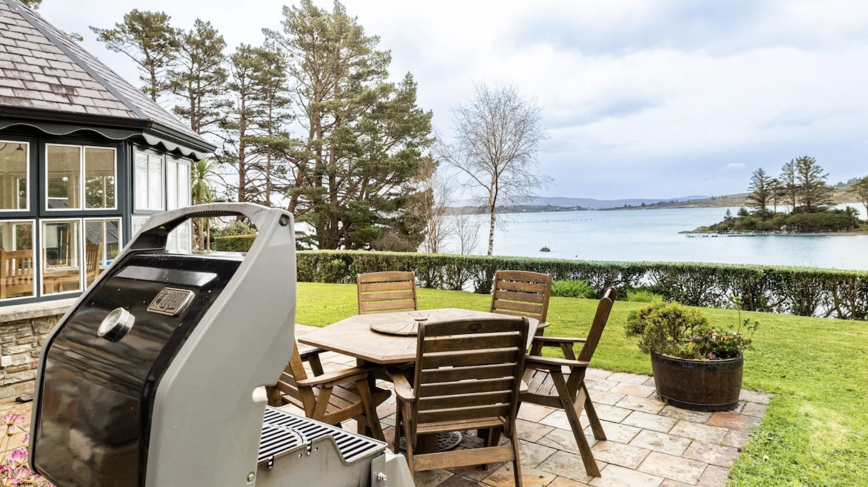 Retreat house in Ireland's patio with wooden dining table and chairs overlooking a lake, with a barbecue grill in the foreground, surrounded by green grass, hedges, and trees.