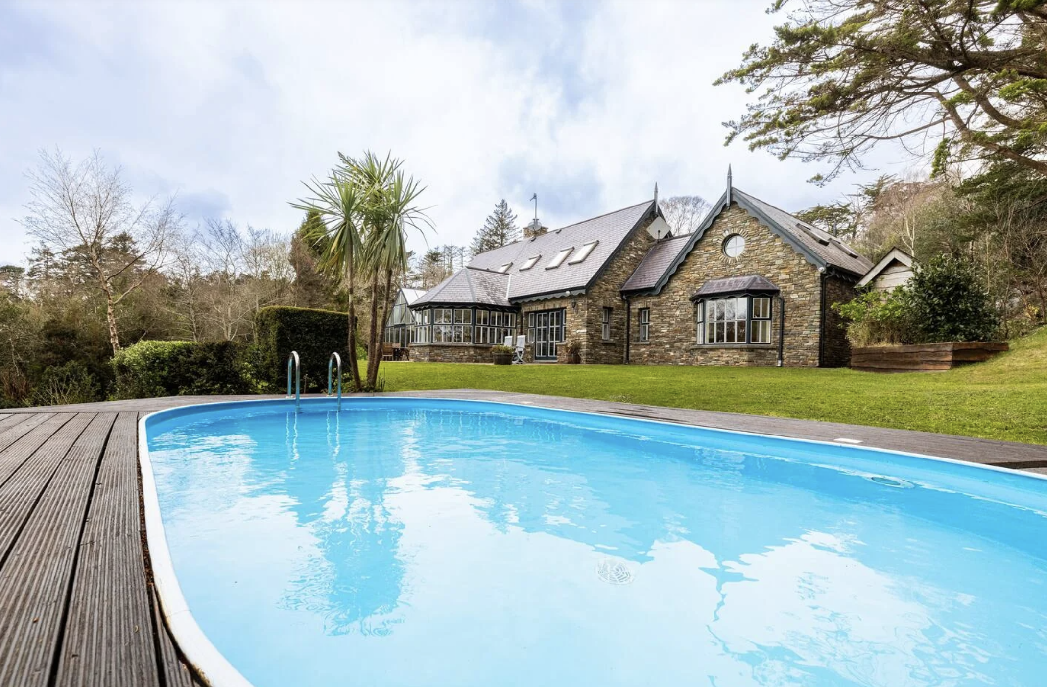 A backyard with a swimming pool, green lawn, tall trees, and a large stone house under a partly cloudy sky at the location of a yoga and art retreat in Ireland.