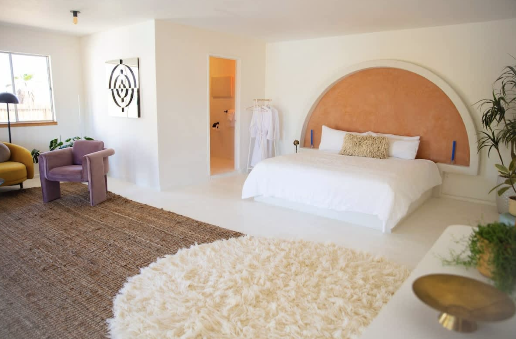 A bedroom with a bed featuring a colorful semi-circular headboard, a white duvet, and a beige pillow. There are plants on the right, a closet with robes to the left, a rug on the floor, and a sitting area with colorful chairs in the background.