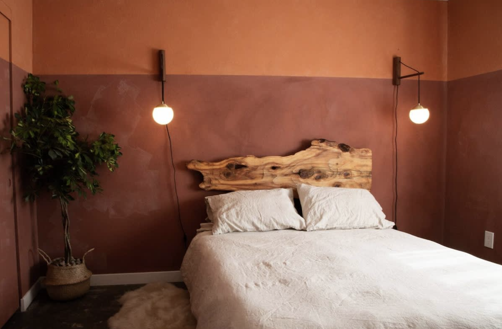 A bedroom with a bed, white bedding, two pillows, a unique wooden headboard, and two hanging wall lamps on a peach-colored wall. There is a large potted plant on the left and a fluffy rug on the floor.