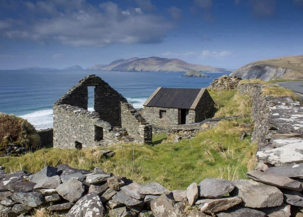 Ruins of an old stone building overlooking the ocean with hills in the background, on a partly cloudy day.