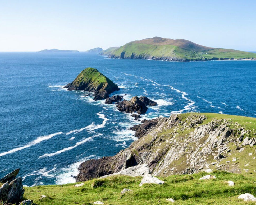 A coastal landscape with green grassy cliffs, rocky islands, and the open ocean under a clear blue sky.