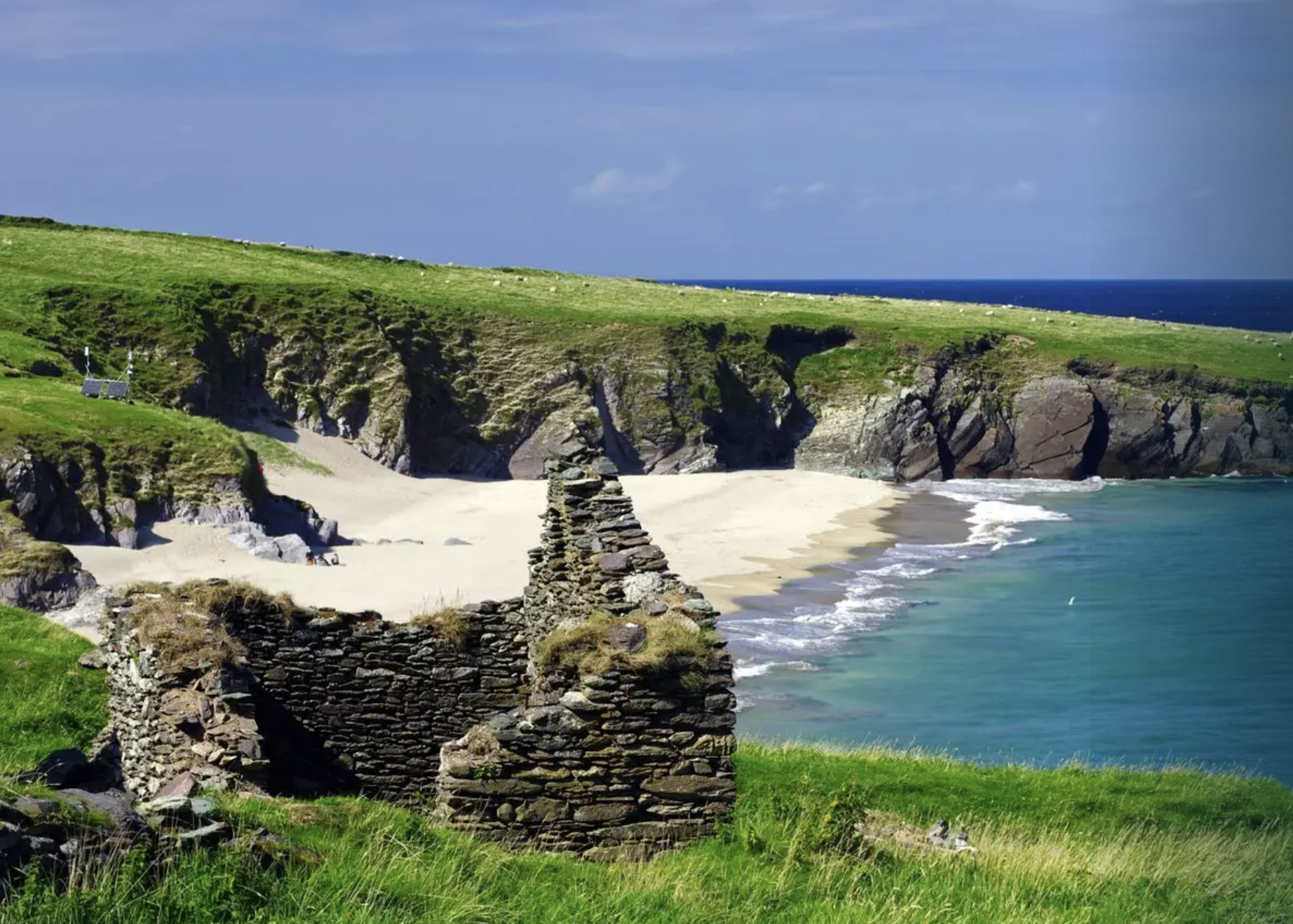 A scenic coastal landscape with green grassy cliffs, a sandy beach, and blue ocean waters, featuring a small stone structure in the foreground.