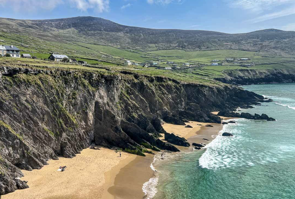 Scenic view of a rocky coastal beach with cliffs, sandy shore, and turquoise water, with houses on a green hillside in the background under a partly cloudy sky.