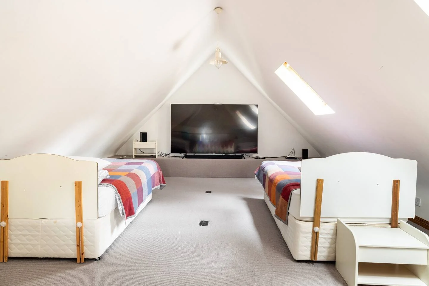 Attic bedroom with two single beds with white headboards and colorful plaid blankets, a large flat-screen TV on a stand, and a skylight window with natural light.